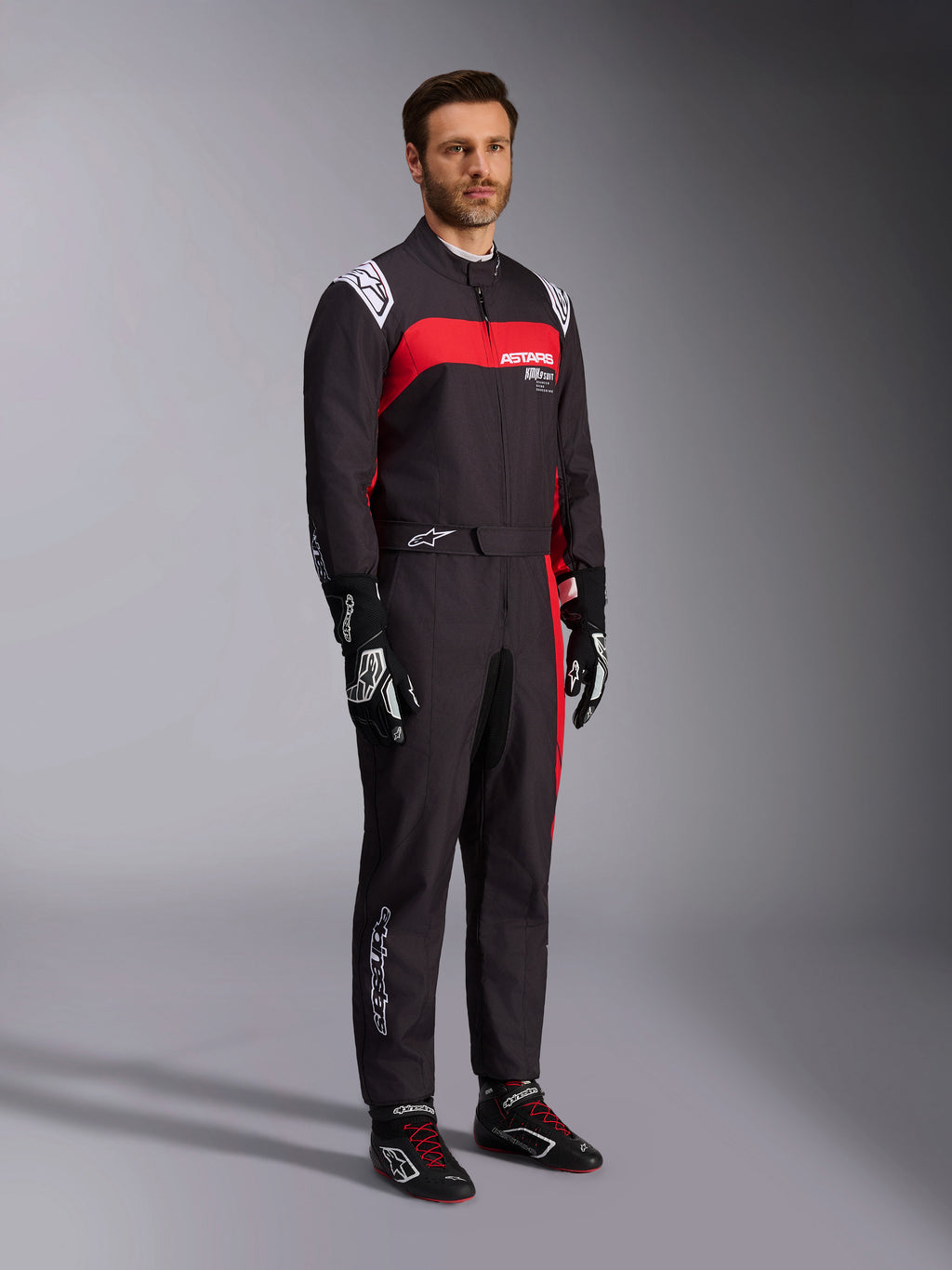 Męski model w kombinezonie Alpinestars KMX-9 V3 Graphic 3, kombinezon kartingowy, czarno-czerwony, z białymi epoletami na ramionach i czerwonym logo na piersi, czarne rękawice wyścigowe i buty z czerwonymi sznurowadłami, profesjonalna odzież motorsportowa.