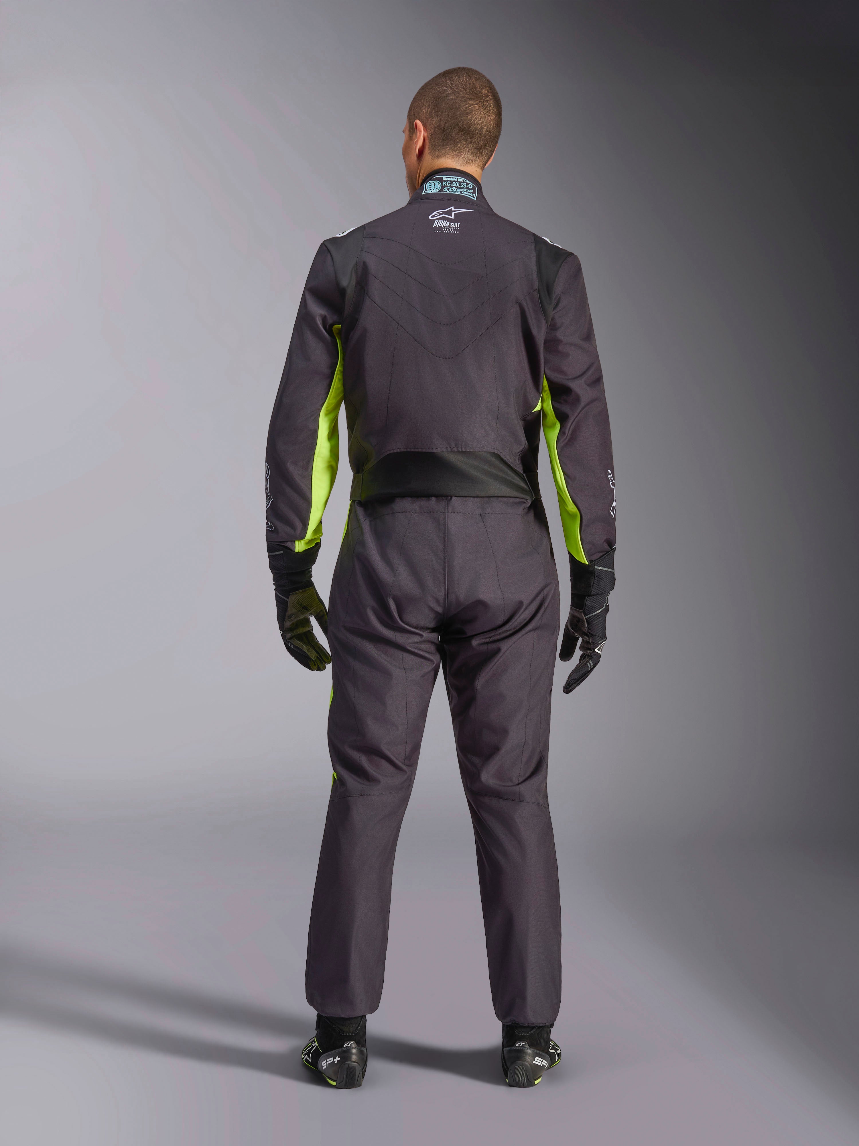 Widok od tyłu na całą sylwetkę osoby ubranej w kombinezon kartingowy Alpinestars KMX-9 V3 Graphic 3 w kolorze czarnym i żółtym fluo, widoczne ergonomiczne panele rozciągliwe na plecach i ramionach, czarne rękawice i buty wyścigowe.