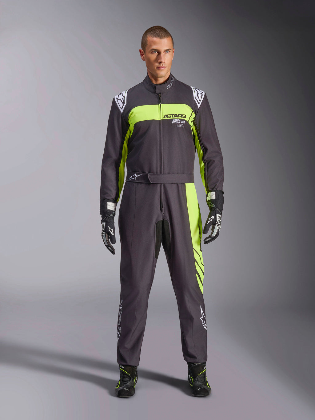 Alpinestars KMX-9 V3 Graphic 3 Suit, czarno-żółty fluo, profesjonalny jednoczęściowy kombinezon kartingowy prezentowany przez modela, z wyraźnymi neonowymi żółtymi akcentami, logo ASTARS na klatce piersiowej oraz pasującymi rękawicami i butami wyścigowymi.