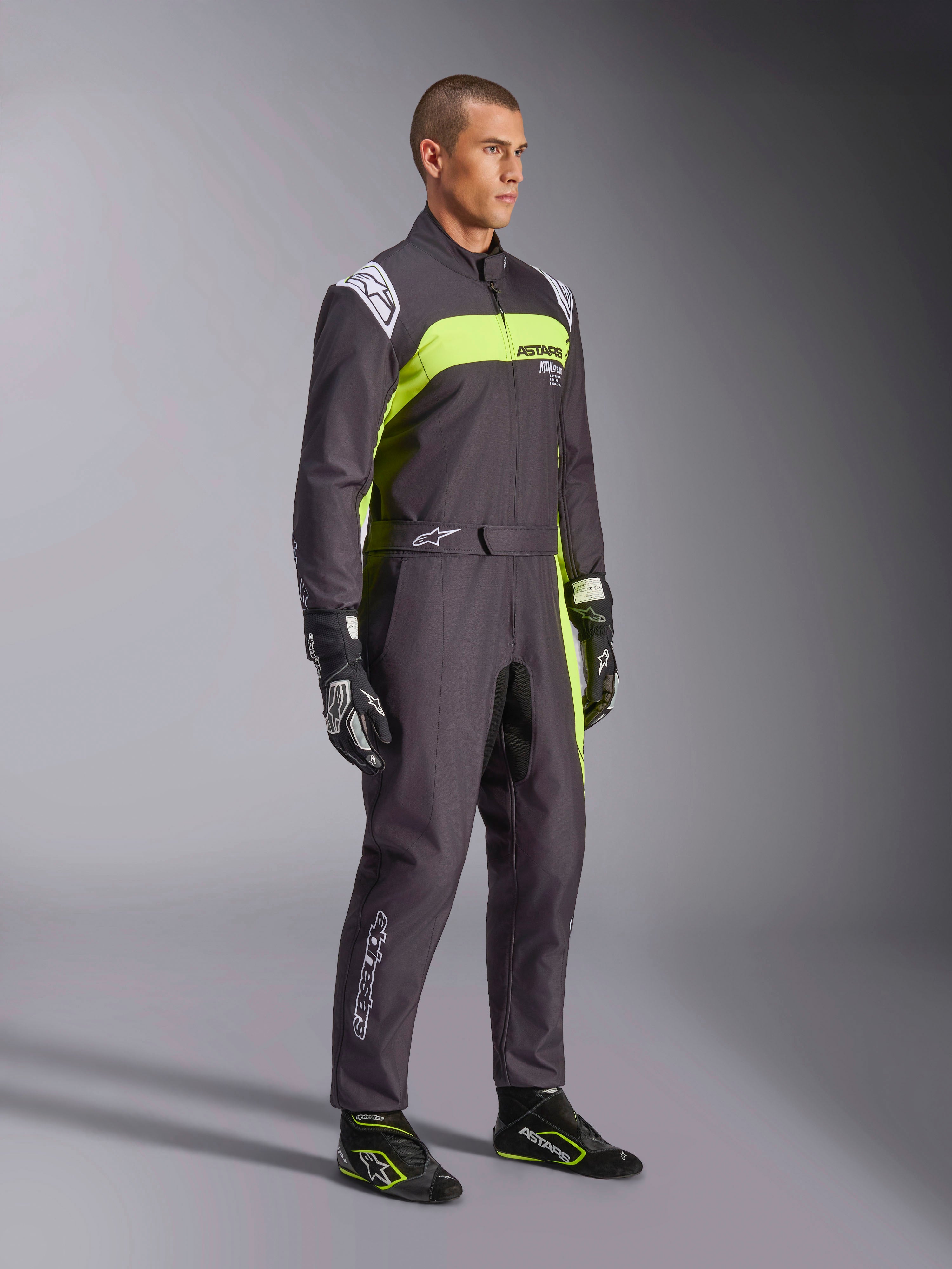 Męski model w kombinezonie Alpinestars KMX-9 V3 Graphic 3 w kolorze czarnym i żółtym fluorescencyjnym, profesjonalny strój kartingowy z jaskrawymi neonowymi żółtymi panelami i białymi akcentami logo, z dopasowanymi rękawicami i butami wyścigowymi, na tle studyjnym.