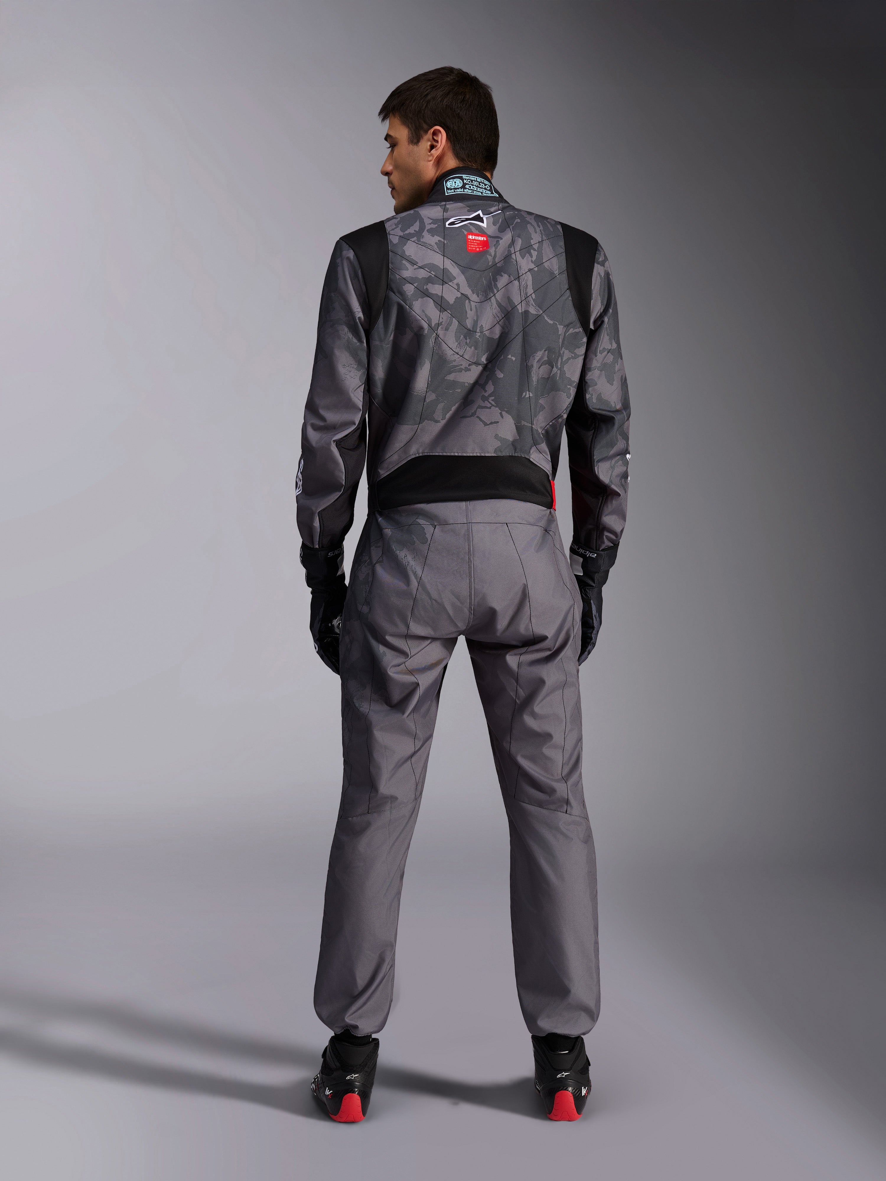 Alpinestars KMX-9 V3 Graphic 5 Suit, Karting Suit, Dark Gray and Black camouflage pattern on the upper body, back view showing stretch panels and Alpinestars branding, worn with black gloves and black racing shoes with red details, professional motorsport racing gear