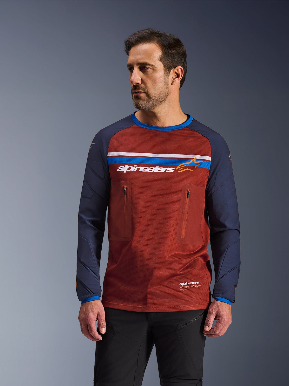 Męski model w koszulce Alpinestars Maxdura Dual Jersey, rdzawobrązowa i granatowa koszulka motocyklowa adventure, dwie kieszenie na piersi na zamek, poziomy pas z logo, korpus z technicznej siatki i wytrzymałe tkane rękawy.