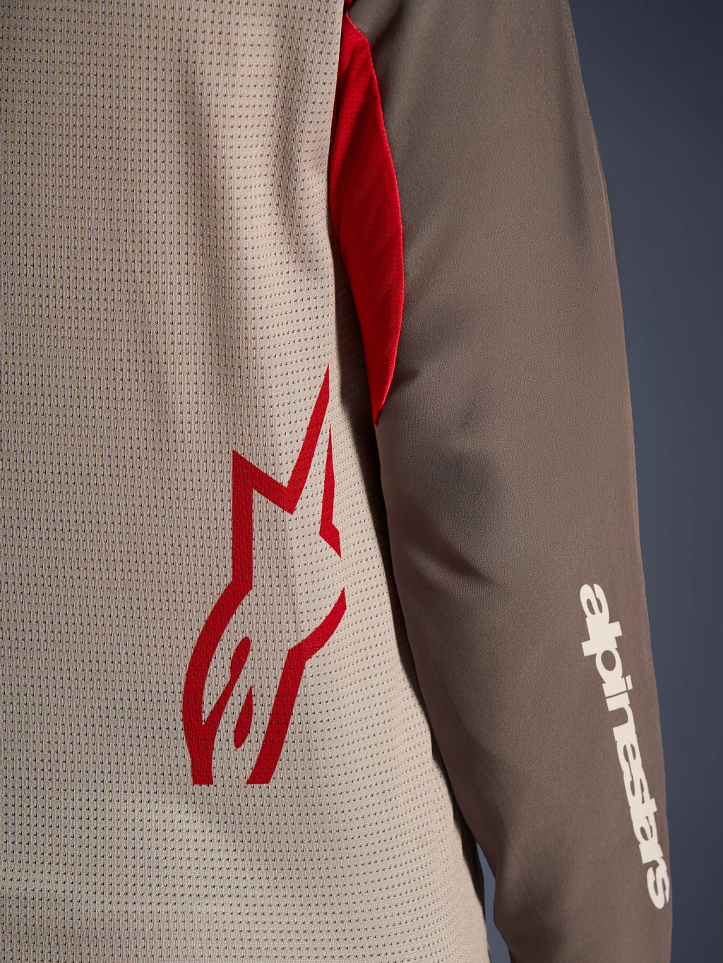 Alpinestars Maxdura Dual Jersey, Aluminum Walnut, zbliżenie na wentylowany siatkowy tors z czerwonym logo gwiazdy i brązowy rękaw odporny na ścieranie z białym napisem Alpinestars, techniczna odzież motocyklowa adventure i dual-sport