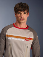 Męski model w koszulce Alpinestars Maxdura Dual Jersey w kolorze Aluminum Walnut, przygodowa koszulka motocyklowa z korpusem z pełnej siatki, odporne na ścieranie rękawy, czerwony kołnierzyk, kieszenie na klatce piersiowej na zamek, pomarańczowe i białe pasy z logo Alpinestars