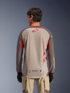 Widok od tyłu na mężczyznę w koszulce Alpinestars Maxdura Dual Jersey w kolorze Aluminum Walnut, koszulka motocyklowa typu adventure z beżowym korpusem z siateczki i czerwonym logo gwiazdy, ciemnobrązowe rękawy z czerwonymi akcentami, beżowe spodnie motocyklowe i czarne rękawice, zaprojektowana dla wysokiej wydajności wentylacji off-road.