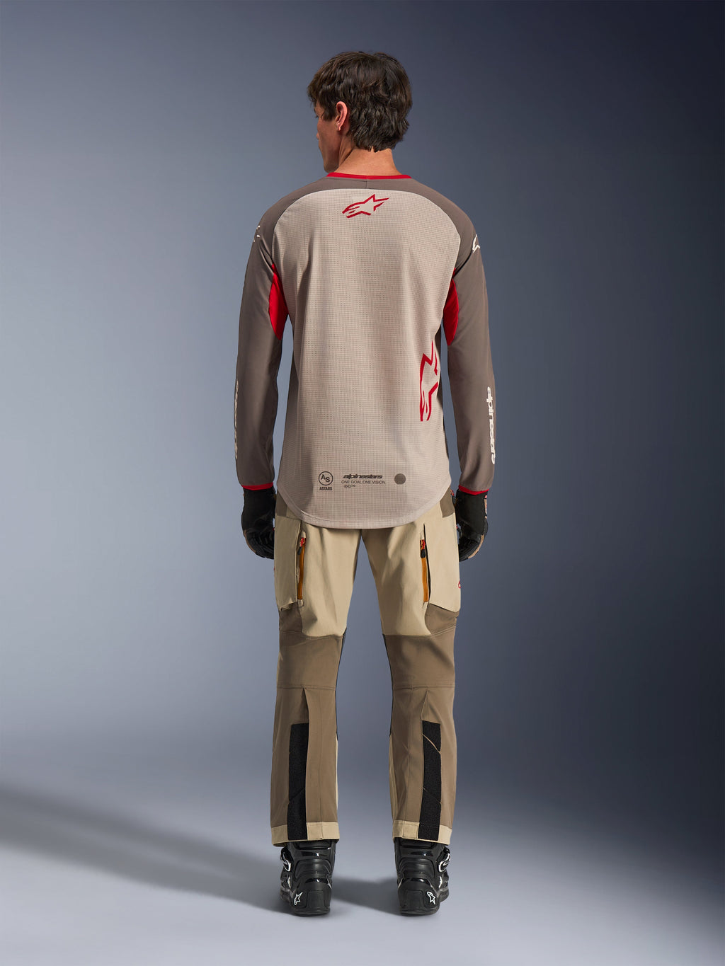Koszulka Alpinestars Maxdura Dual w kolorze Aluminum Walnut, widok z tyłu, beżowy tułów z siatki z brązowymi rękawami i czerwonymi logotypami, wydłużony tył, w zestawieniu ze spodniami adventure i czarnymi butami.