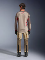 Koszulka Alpinestars Maxdura Dual w kolorze Aluminum Walnut, widok z tyłu, beżowy tułów z siatki z brązowymi rękawami i czerwonymi logotypami, wydłużony tył, w zestawieniu ze spodniami adventure i czarnymi butami.