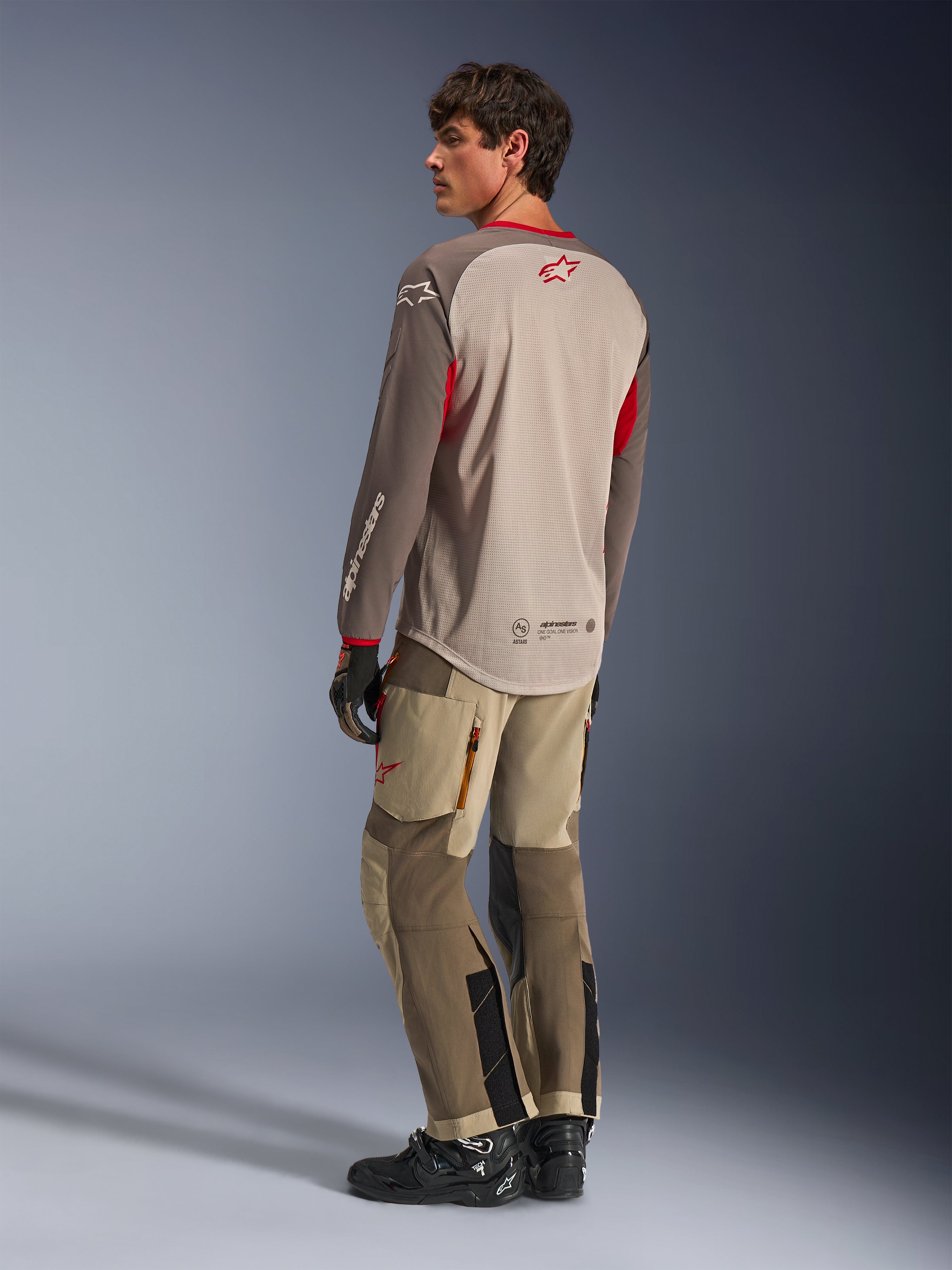 Alpinestars Maxdura Dual Jersey w kolorze Aluminum Walnut, widok z tyłu motocyklowej koszulki adventure z oddychającym siatkowym torsem i czerwonymi akcentami, zestawiona z pasującymi technicznymi spodniami adventure i czarnymi butami