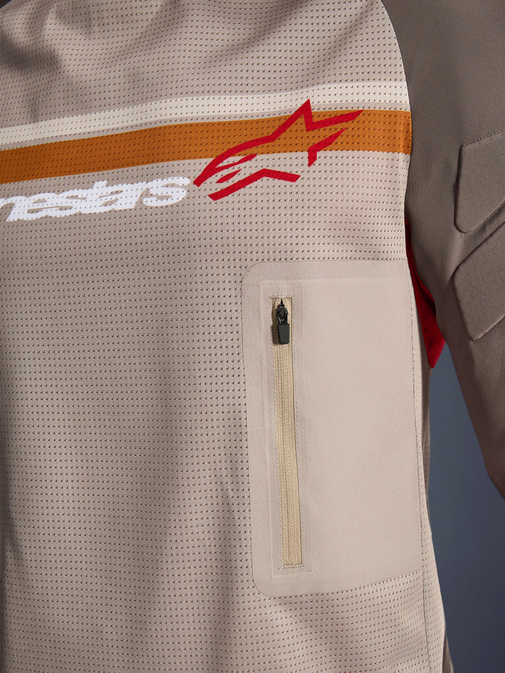 Alpinestars Maxdura Dual Jersey, Aluminum Walnut, zbliżenie na detale siatkowego korpusu, poziomy pomarańczowo-biały pas z czerwonym logo, pionowa kieszeń na piersi na zamek, odzież motocyklowa typu adventure