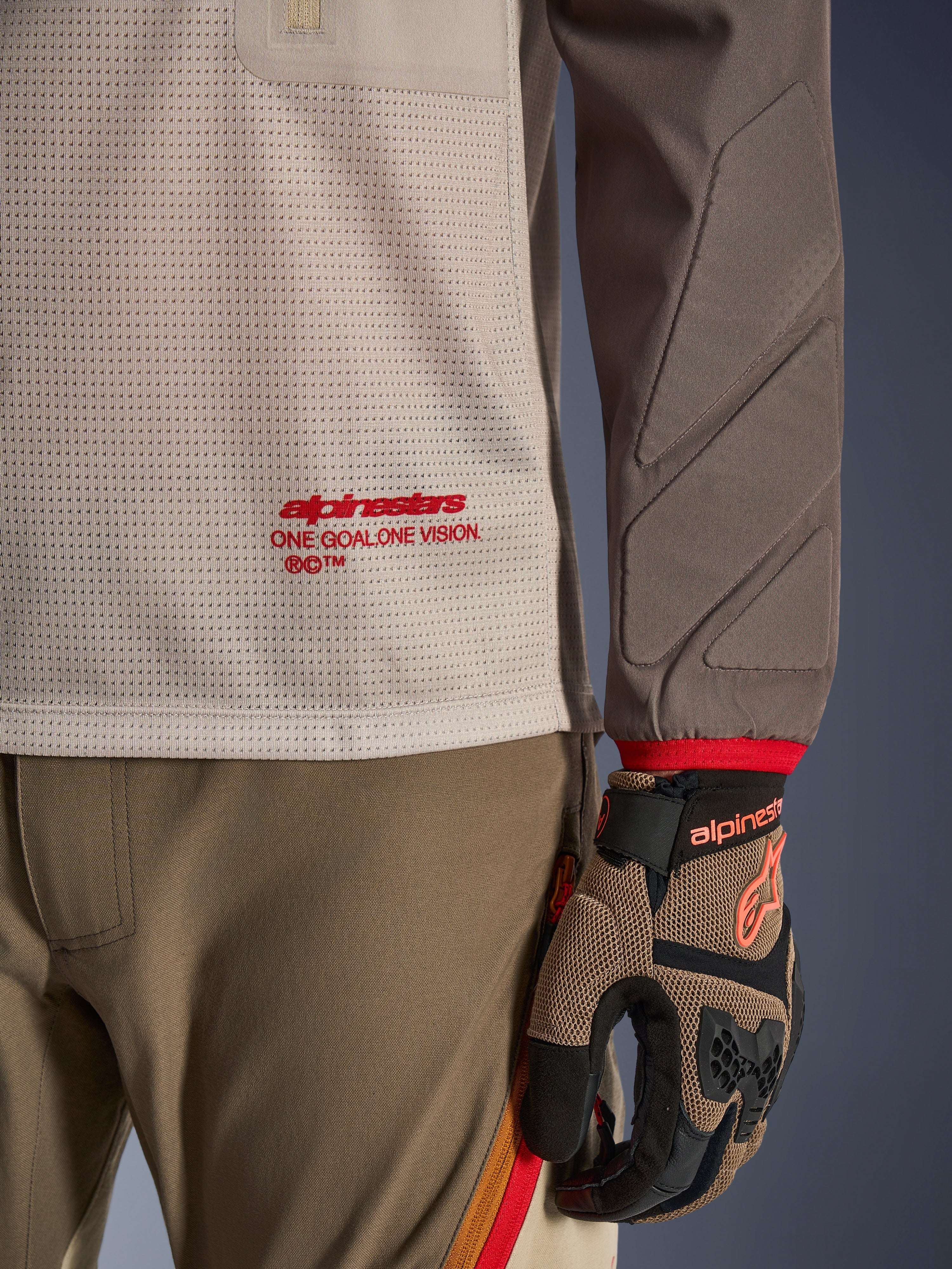 Alpinestars Maxdura Dual Jersey w kolorze Aluminum Walnut, widok szczegółowy pokazujący wentylowany siatkowy tors z czerwonym logo, rękaw odporny na ścieranie ze zintegrowanymi wyściółkami, noszona z dopasowanymi spodniami i rękawicami adventure