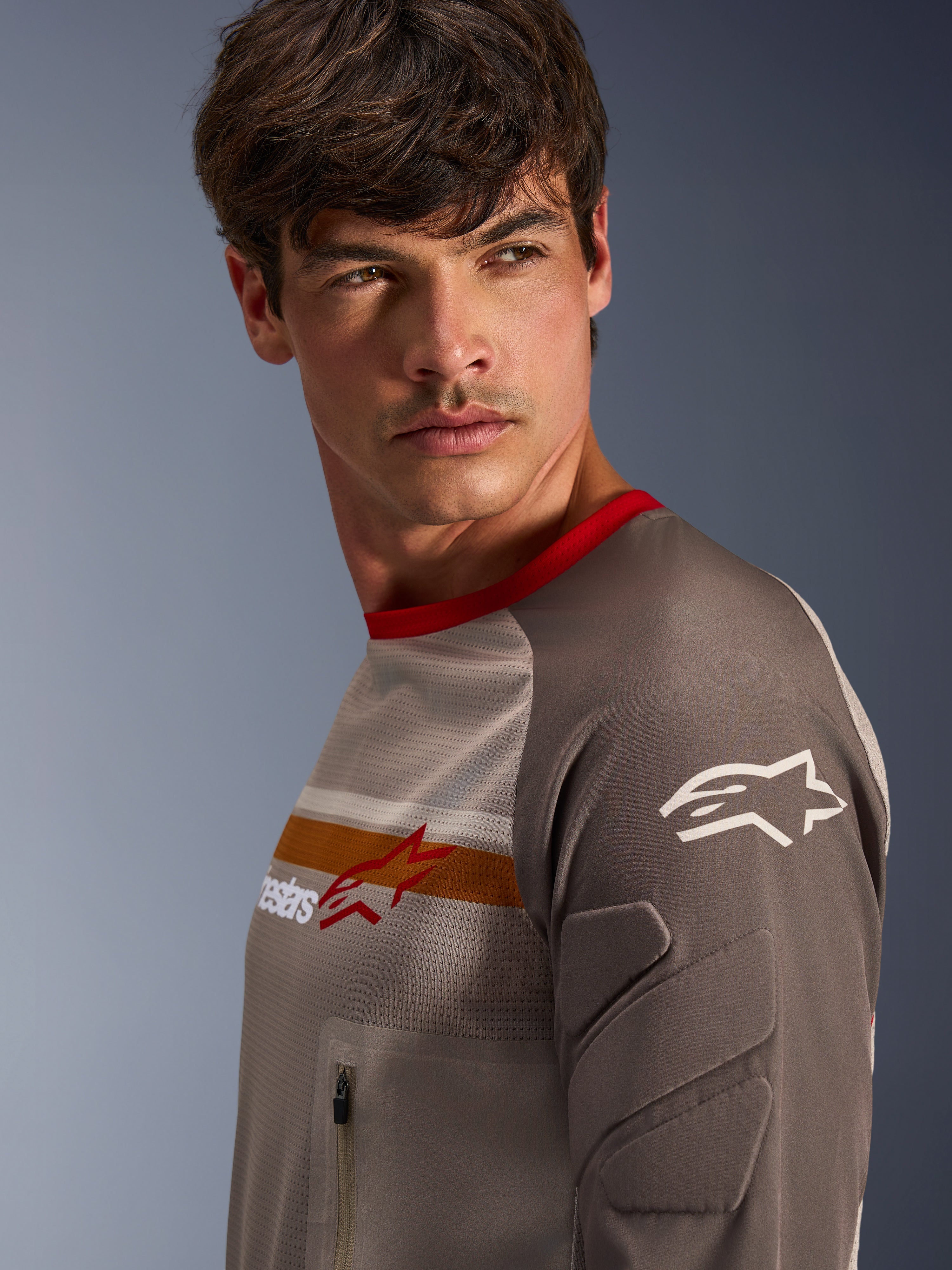 Męski model w koszulce Alpinestars Maxdura Dual Jersey w kolorze Aluminum Walnut, z wentylowanym siatkowym torsem, czerwonym kołnierzykiem i pomarańczowym pasem na klatce piersiowej. Zbliżenie na odporny na ścieranie rękaw ze zintegrowanymi wyściółkami oraz zapinaną na zamek kieszeń na klatce piersiowej do jazdy adventure.