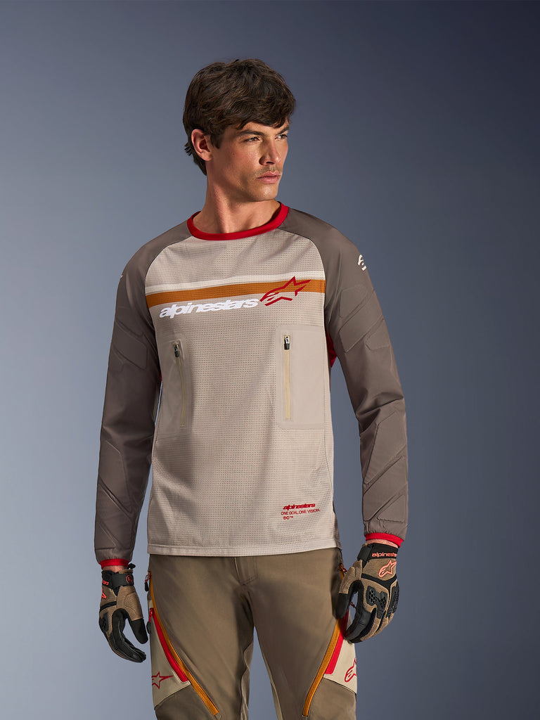 Alpinestars Maxdura Dual Jersey w kolorze Aluminum Walnut, przygodowa koszulka motocyklowa z oddychającym siateczkowym korpusem, odpornymi na ścieranie rękawami w odcieniu walnut brown i zapinanymi kieszeniami na piersi, noszona przez modela z dopasowanym ekwipunkiem terenowym.