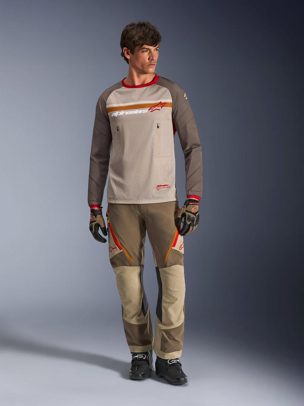Alpinestars Maxdura Dual Jersey w kolorze Aluminum Walnut, koszulka motocyklowa adventure z beżowym siatkowym korpusem i brązowymi rękawami, z czerwonym kołnierzykiem i poziomym pasem na klatce piersiowej z logo Alpinestars, noszona z dopasowanymi spodniami adventure i rękawicami.
