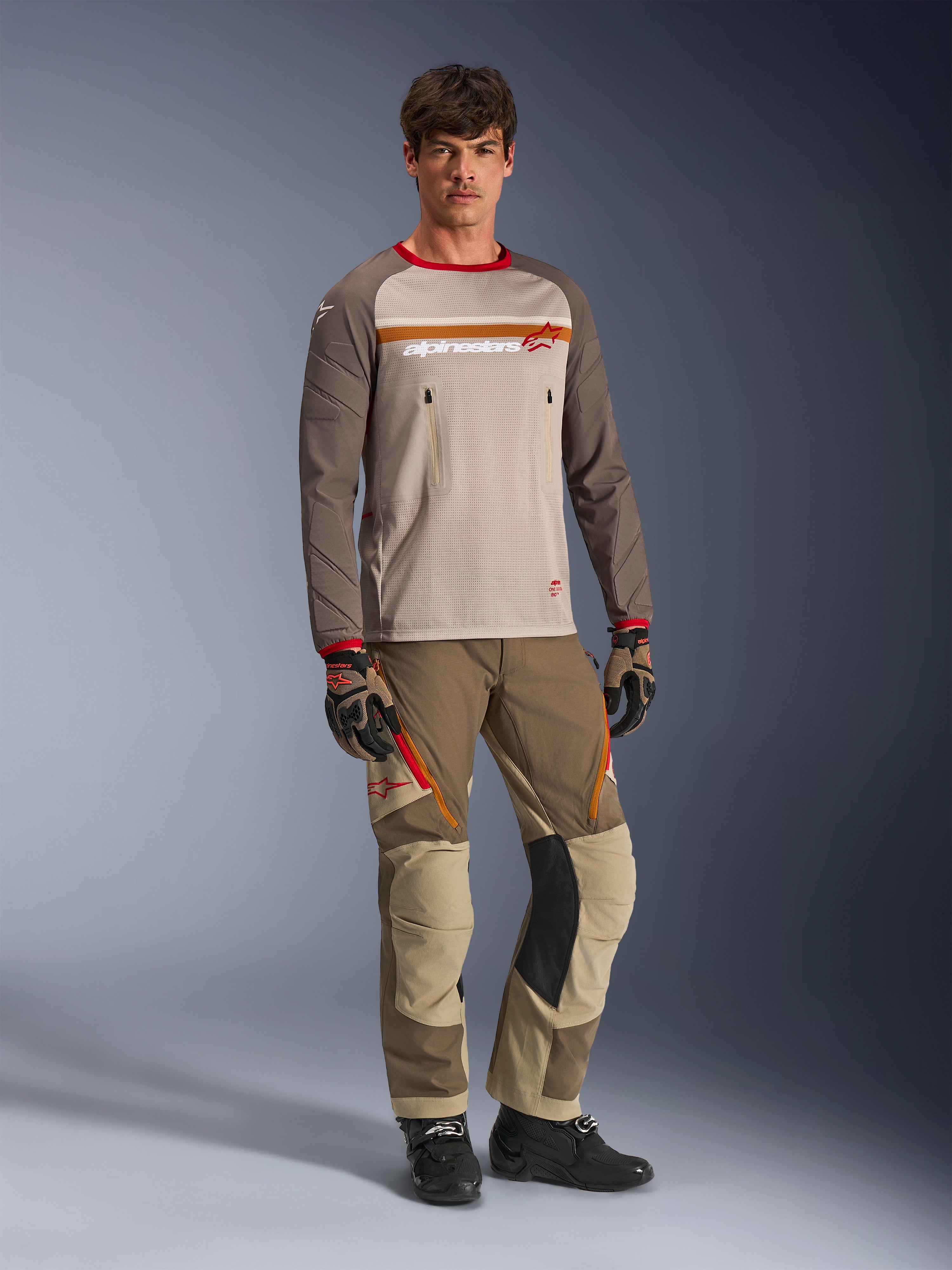 Model w koszulce Alpinestars Maxdura Dual Jersey w kolorze Aluminum Walnut, z szarym siatkowym torsem, brązowymi rękawami z czerwonymi mankietami i kieszeniami na klatce piersiowej na zamek, w stylizacji z pasującym sprzętem terenowym do jazdy adventure.