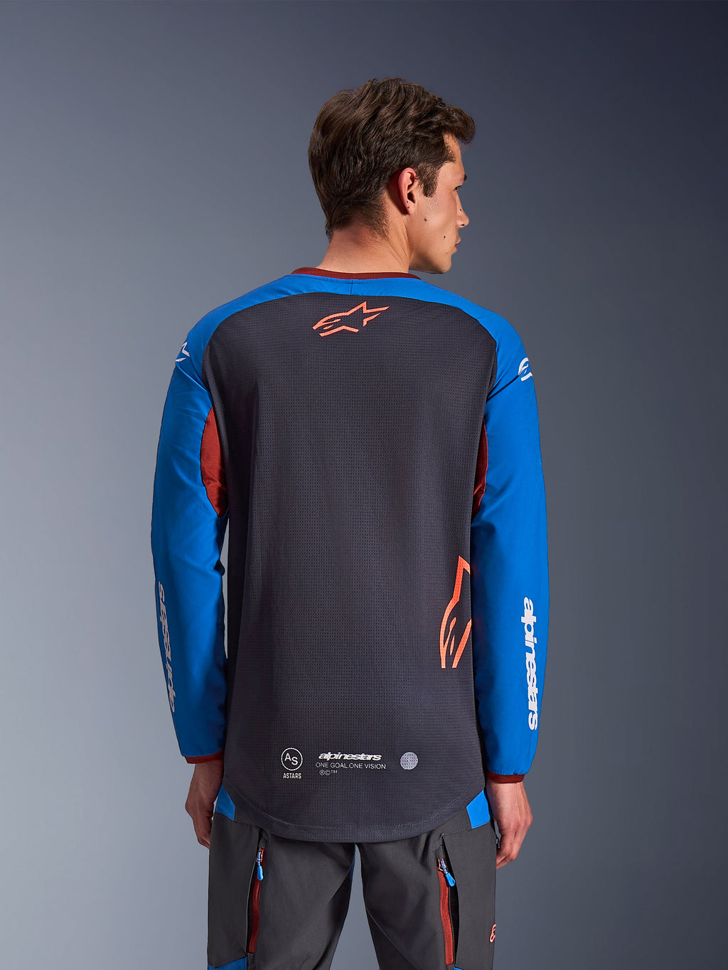 Alpinestars Maxdura Dual Jersey w kolorze ciemnoszarym i królewskim niebieskim, widok od tyłu na modelu, siateczkowa konstrukcja tułowia z pomarańczowym logo Astars, długie rękawy z białym brandingiem, bordowy kołnierzyk i mankiety, zestaw z szarymi spodniami technicznymi, odzież motocyklowa dual-sport i adventure