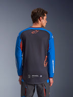 Alpinestars Maxdura Dual Jersey w kolorze ciemnoszarym i królewskim niebieskim, widok od tyłu na modelu, siateczkowa konstrukcja tułowia z pomarańczowym logo Astars, długie rękawy z białym brandingiem, bordowy kołnierzyk i mankiety, zestaw z szarymi spodniami technicznymi, odzież motocyklowa dual-sport i adventure