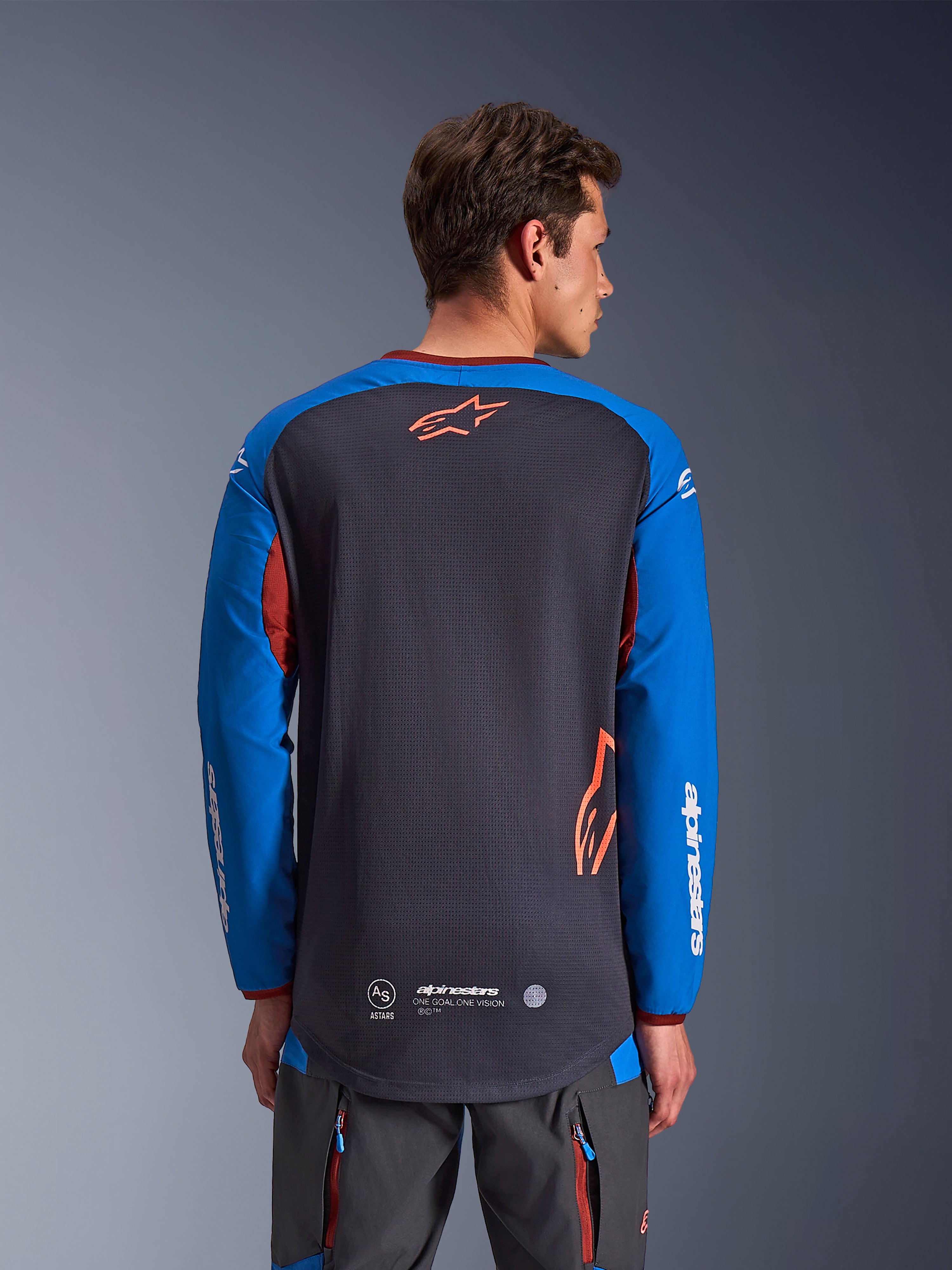 Alpinestars Maxdura Dual Jersey w kolorze ciemnoszarym i królewskim niebieskim, widok od tyłu na modelu, siateczkowa konstrukcja tułowia z pomarańczowym logo Astars, długie rękawy z białym brandingiem, bordowy kołnierzyk i mankiety, zestaw z szarymi spodniami technicznymi, odzież motocyklowa dual-sport i adventure