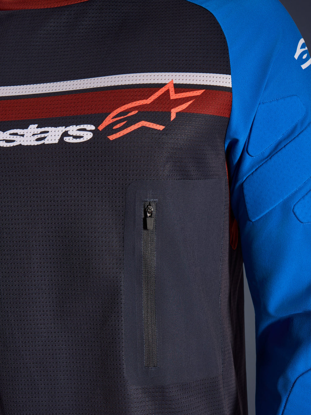 Alpinestars Maxdura Dual Jersey, zbliżenie na klatkę piersiową i rękaw, ciemnoszary i niebieski, zapinana na zamek kieszeń na piersi, oddychający siatkowy tułów, zintegrowane ochraniacze na rękawach, logo Alpinestars na tle czerwonych i białych pasów