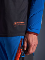 Zbliżenie na koszulkę Alpinestars Maxdura Dual w kolorze ciemnoszarym i niebieskim, pomarańczowe logo i napis „One Goal One Vision” na siatkowym tułowiu, wyściełany rękaw ze wzmocnionymi panelami geometrycznymi, detal technicznej odzieży adventure