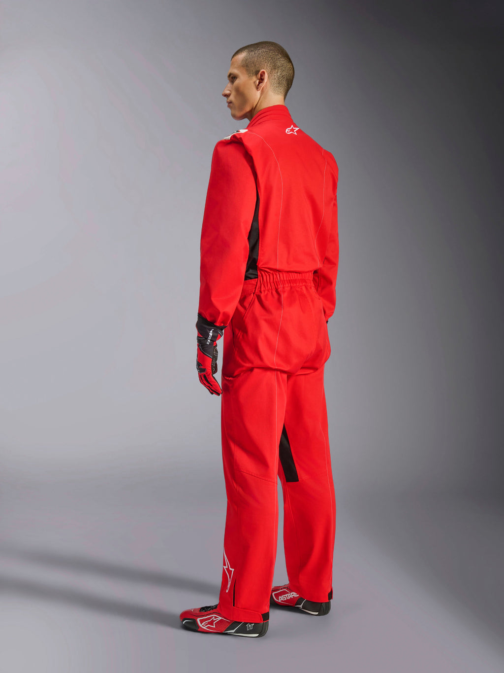 Alpinestars Kart Indoor V2 Suit, czerwony kombinezon kartingowy, widok z tyłu pod kątem trzech czwartych na modelu, zestawiony z pasującymi czerwonymi rękawicami i butami wyścigowymi, jednowarstwowa konstrukcja z kepru polibawełnianego, profesjonalny sprzęt motorsportowy do kartingu halowego.