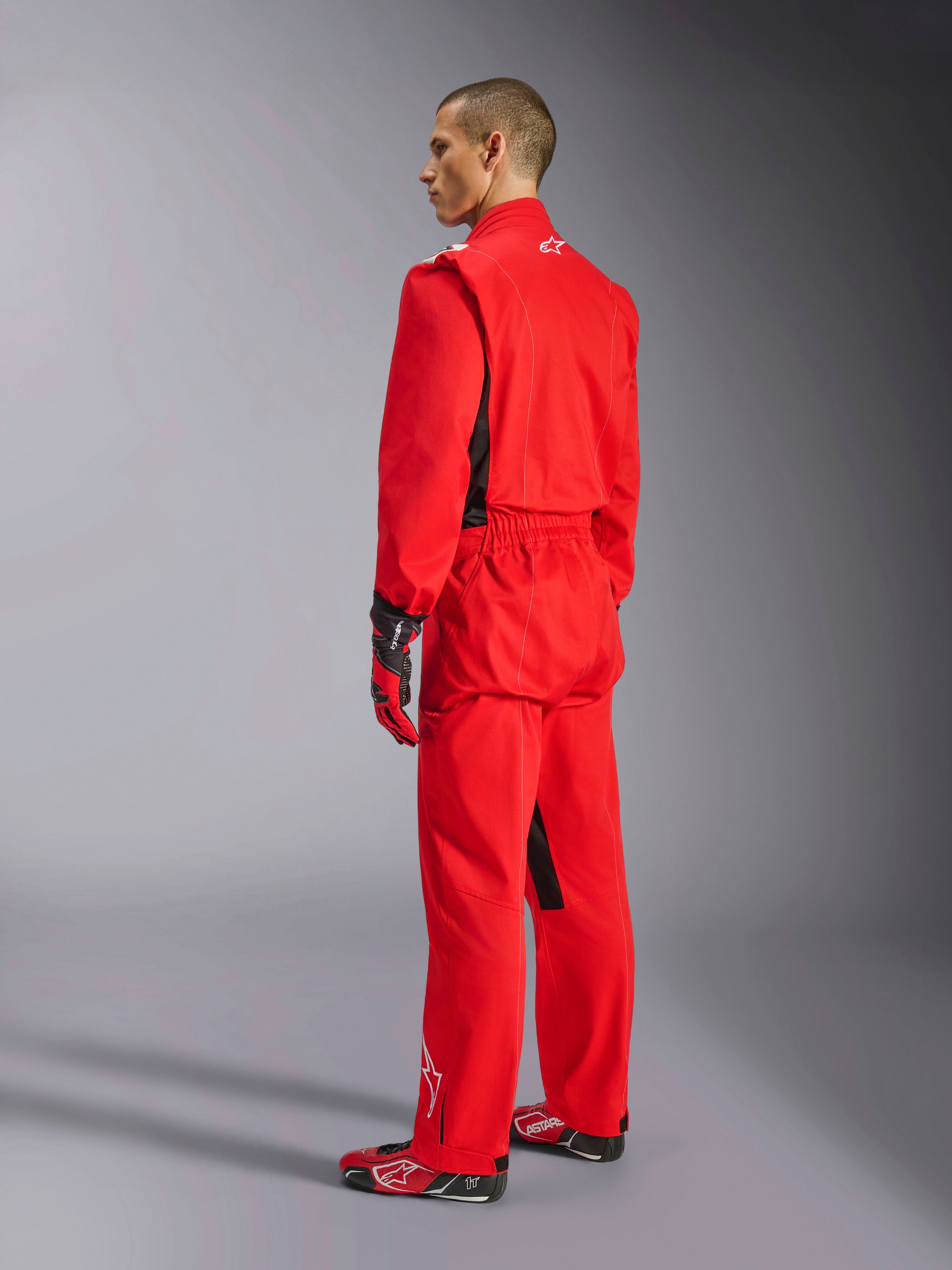 Alpinestars Kart Indoor V2 Suit, czerwony kombinezon kartingowy, widok z tyłu pod kątem trzech czwartych na modelu, zestawiony z pasującymi czerwonymi rękawicami i butami wyścigowymi, jednowarstwowa konstrukcja z kepru polibawełnianego, profesjonalny sprzęt motorsportowy do kartingu halowego.