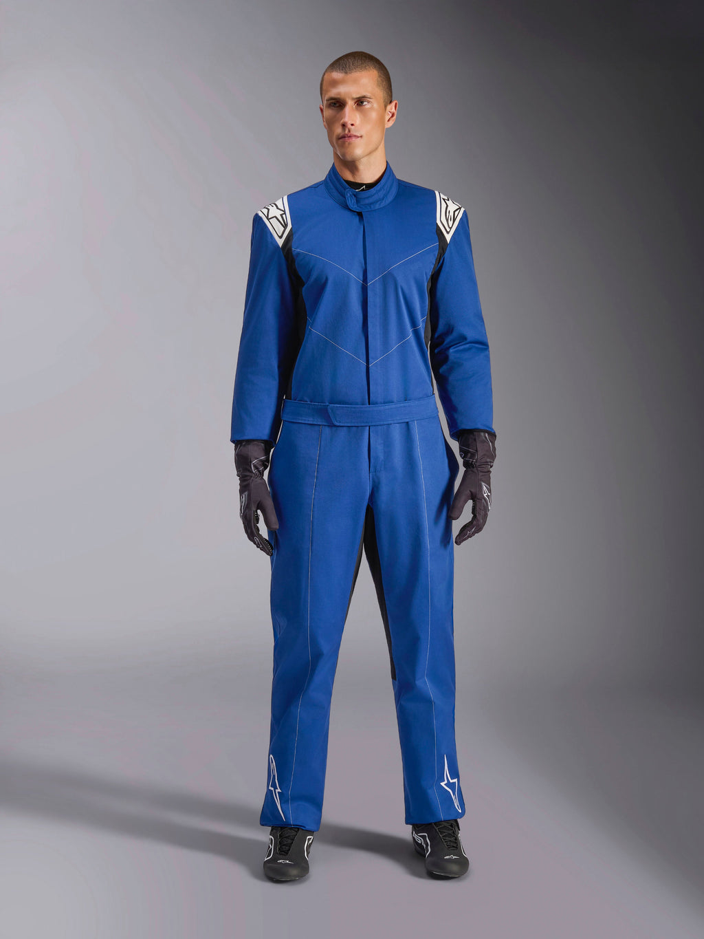 Męski model ubrany w kombinezon Alpinestars Kart Indoor V2 w kolorze niebieskim, jednoczęściowy kombinezon kartingowy z białymi logo gwiazdy na ramionach i goleniach, białe kontrastowe przeszycia, elastyczny pas w talii, zestawiony z czarnymi rękawicami wyścigowymi i obuwiem, zaprojektowany do rekreacyjnego użytku na torze