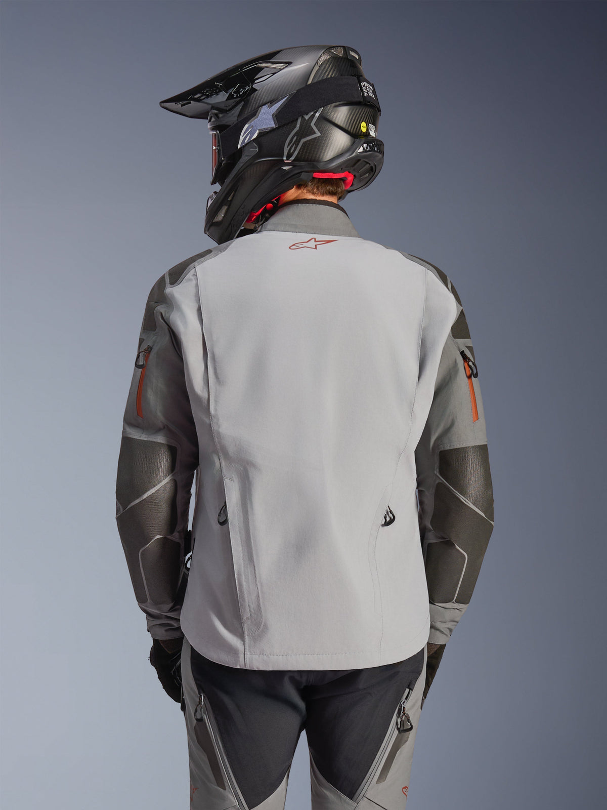 Kurtka Alpinestars Nazca 3L GORE-TEX PRO, motocyklowa kurtka adventure, widok z tyłu, kolor ciemnoszary i średnioszary, posiada wzmocnienia Superfabric na łokciach i ramionach, czerwone logo na plecach, noszona z pasującą odzieżą adventure i kaskiem z włókna węglowego.