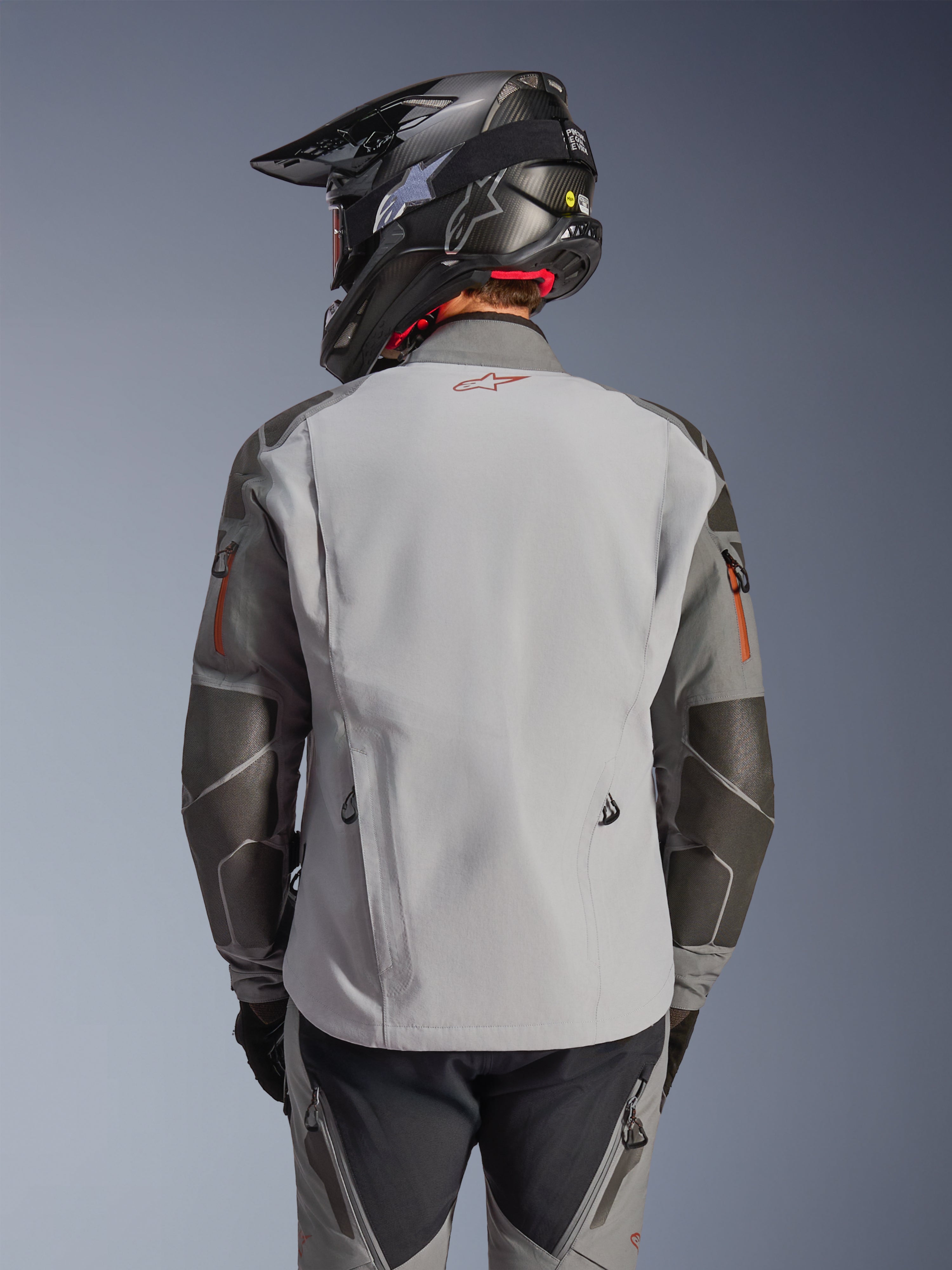 Kurtka Alpinestars Nazca 3L GORE-TEX PRO, motocyklowa kurtka adventure, widok z tyłu, kolor ciemnoszary i średnioszary, posiada wzmocnienia Superfabric na łokciach i ramionach, czerwone logo na plecach, noszona z pasującą odzieżą adventure i kaskiem z włókna węglowego.