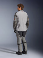 Kurtka Alpinestars Nazca 3L GORE-TEX PRO, motocyklowa kurtka adventure, ciemnoszara i średnioszara, widok z tyłu, noszona z pasującymi spodniami adventure i czarnymi butami, ciemnoszare wzmocnienia Superfabric na rękawach, pomarańczowe akcenty przy zamkach, małe czerwone logo na kołnierzu z tyłu.