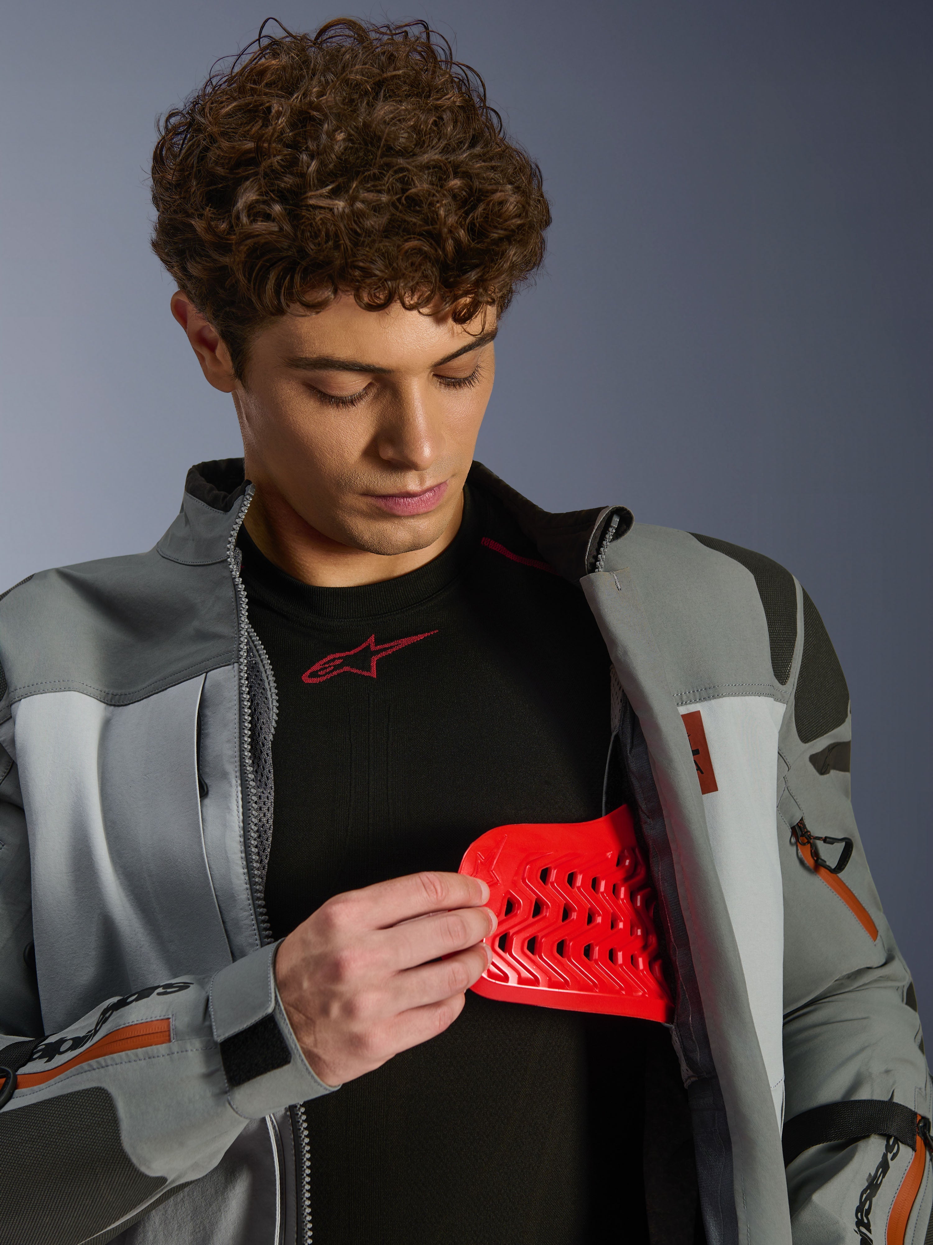 Model w kurtce Alpinestars Nazca 3L GORE-TEX PRO, ciemnoszara i średnioszara, prezentujący czerwoną wkładkę ochronną Nucleon PLASMA Pro, odzież motocyklowa adventure, kurtka premium z technicznymi detalami