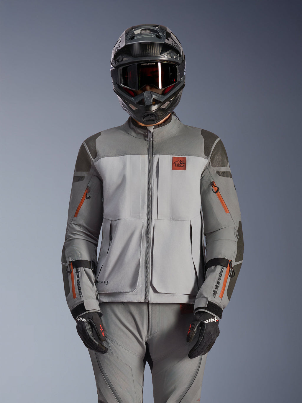 Kurtka Alpinestars Nazca 3L GORE-TEX PRO, motocyklowa kurtka adventure, ciemnoszara i średnioszara z pomarańczowymi akcentami przy zamkach, noszona z pasującymi spodniami i czarnym kaskiem, wytrzymała konstrukcja tekstylna z widocznym brandingiem GORE-TEX PRO i All Terra