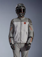 Kurtka Alpinestars Nazca 3L GORE-TEX PRO, motocyklowa kurtka adventure, ciemnoszara i średnioszara z pomarańczowymi akcentami przy zamkach, noszona z pasującymi spodniami i czarnym kaskiem, wytrzymała konstrukcja tekstylna z widocznym brandingiem GORE-TEX PRO i All Terra