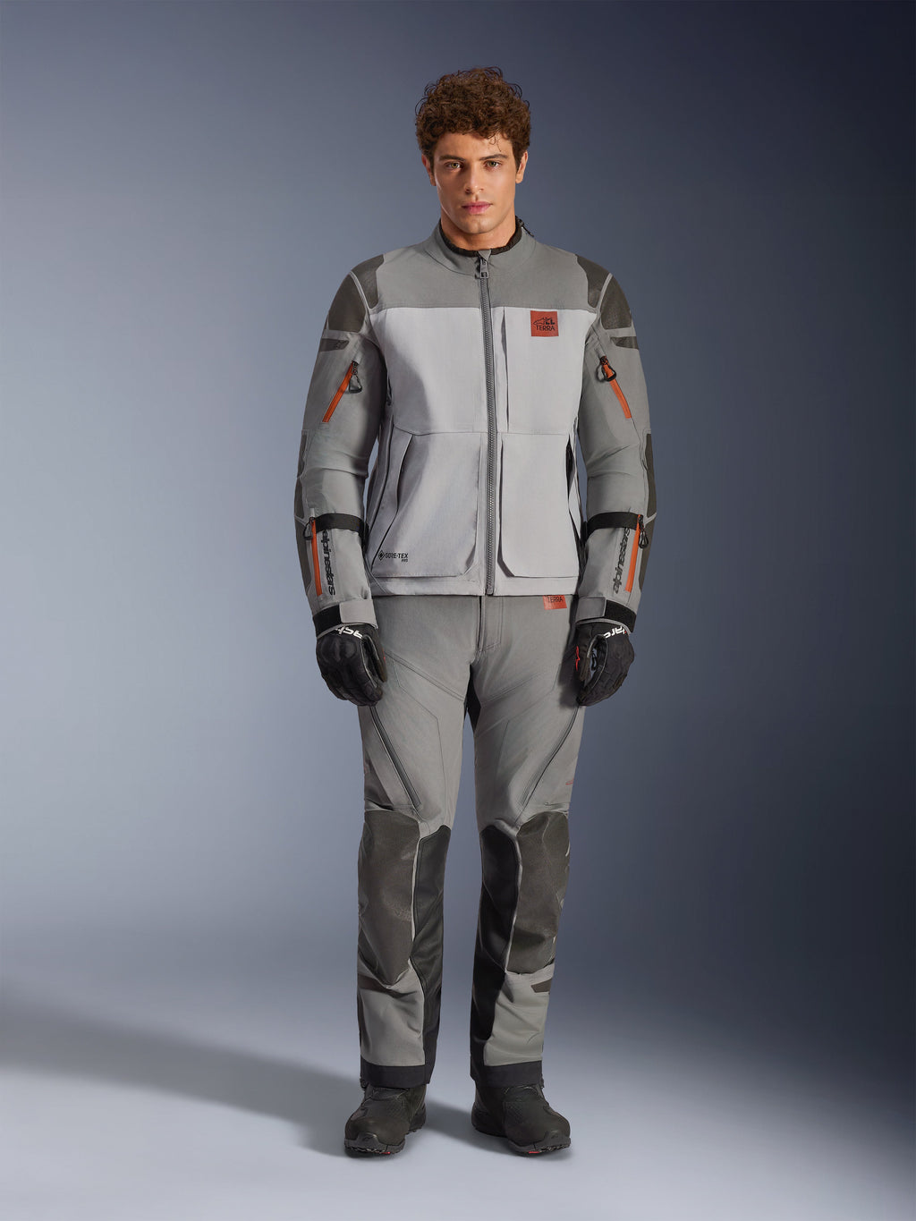 Kurtka Alpinestars Nazca 3L GORE-TEX PRO, motocyklowa kurtka adventure, kolor ciemnoszary i średnioszary, prezentowana przez modela z pasującymi spodniami i czarnymi rękawicami, pomarańczowe akcenty przy zamkach i czerwone logo na piersi, techniczna odzież ochronna do ekstremalnej jazdy
