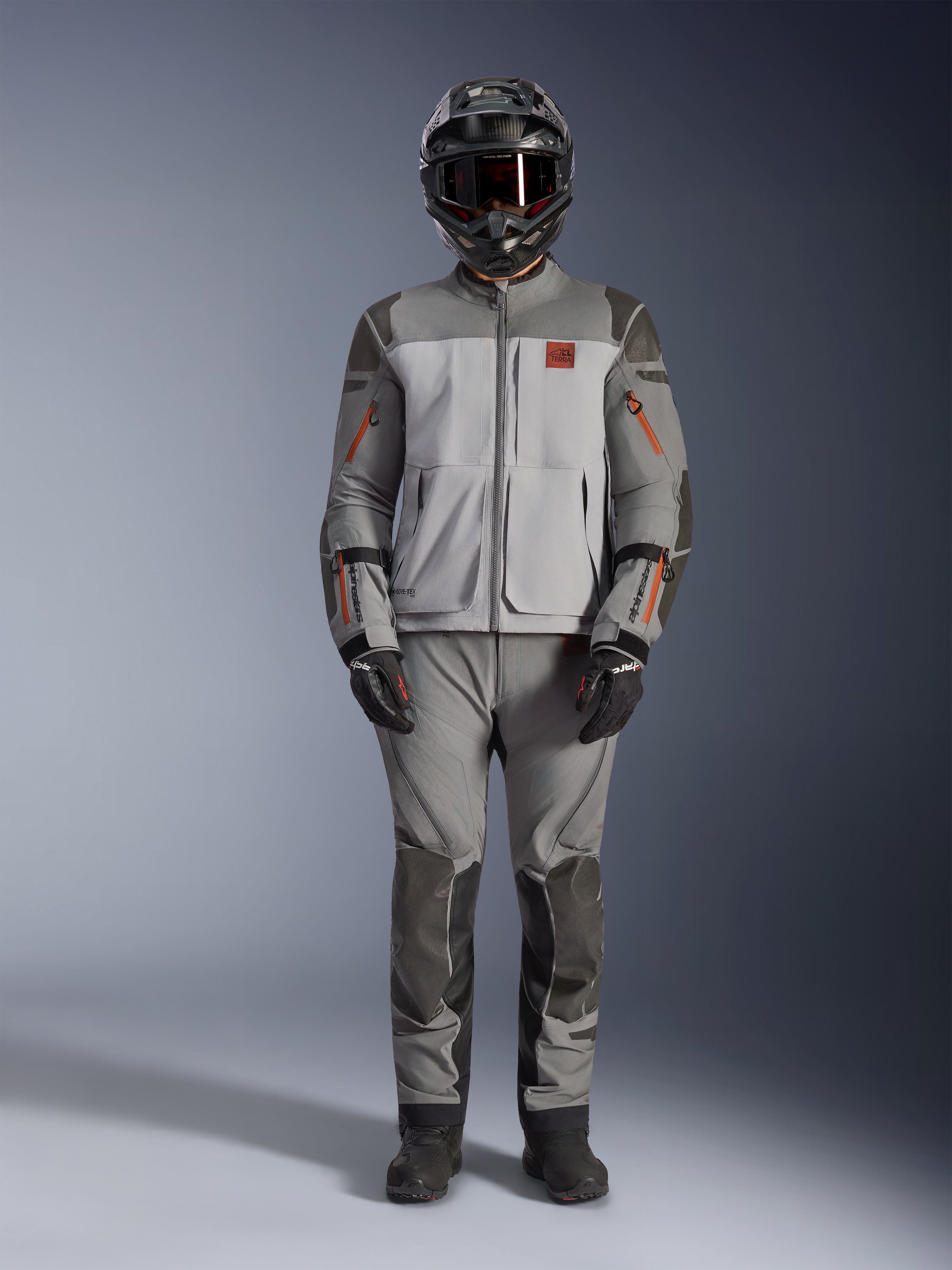 Kurtka Alpinestars Nazca 3L GORE-TEX PRO, motocyklowa kurtka adventure, ciemnoszara i szara z pomarańczowymi akcentami, wzmocnienia Superfabric i przednie zamki wentylacyjne, noszona z pasującymi spodniami i czarnym kaskiem, techniczna odzież terenowa