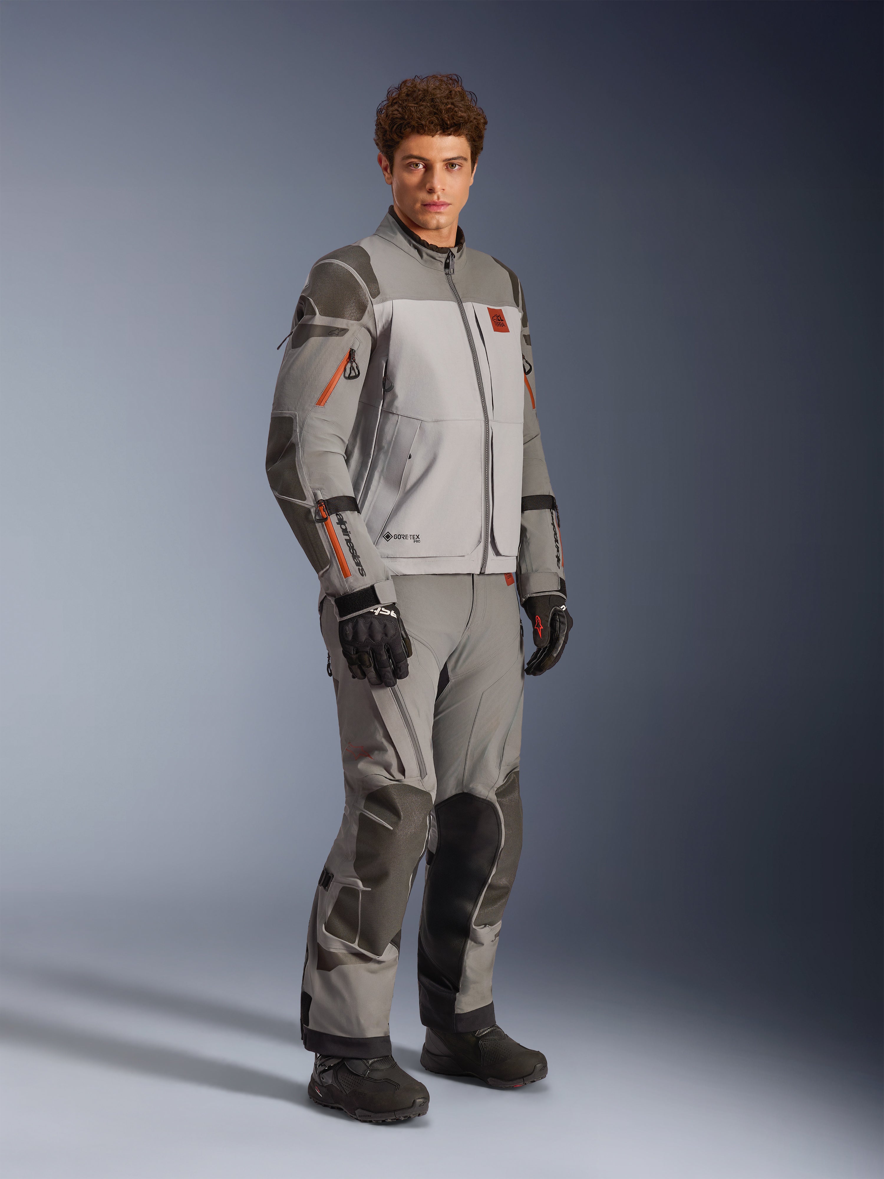 Kurtka Alpinestars Nazca 3L GORE-TEX PRO w kolorze Dark Gray Mid Gray, motocyklowy strój typu adventure noszony z pasującymi spodniami i czarnymi rękawicami, z pomarańczowymi akcentami przy zamkach, ochronnymi wzmocnieniami Superfabric® i logo GORE-TEX.