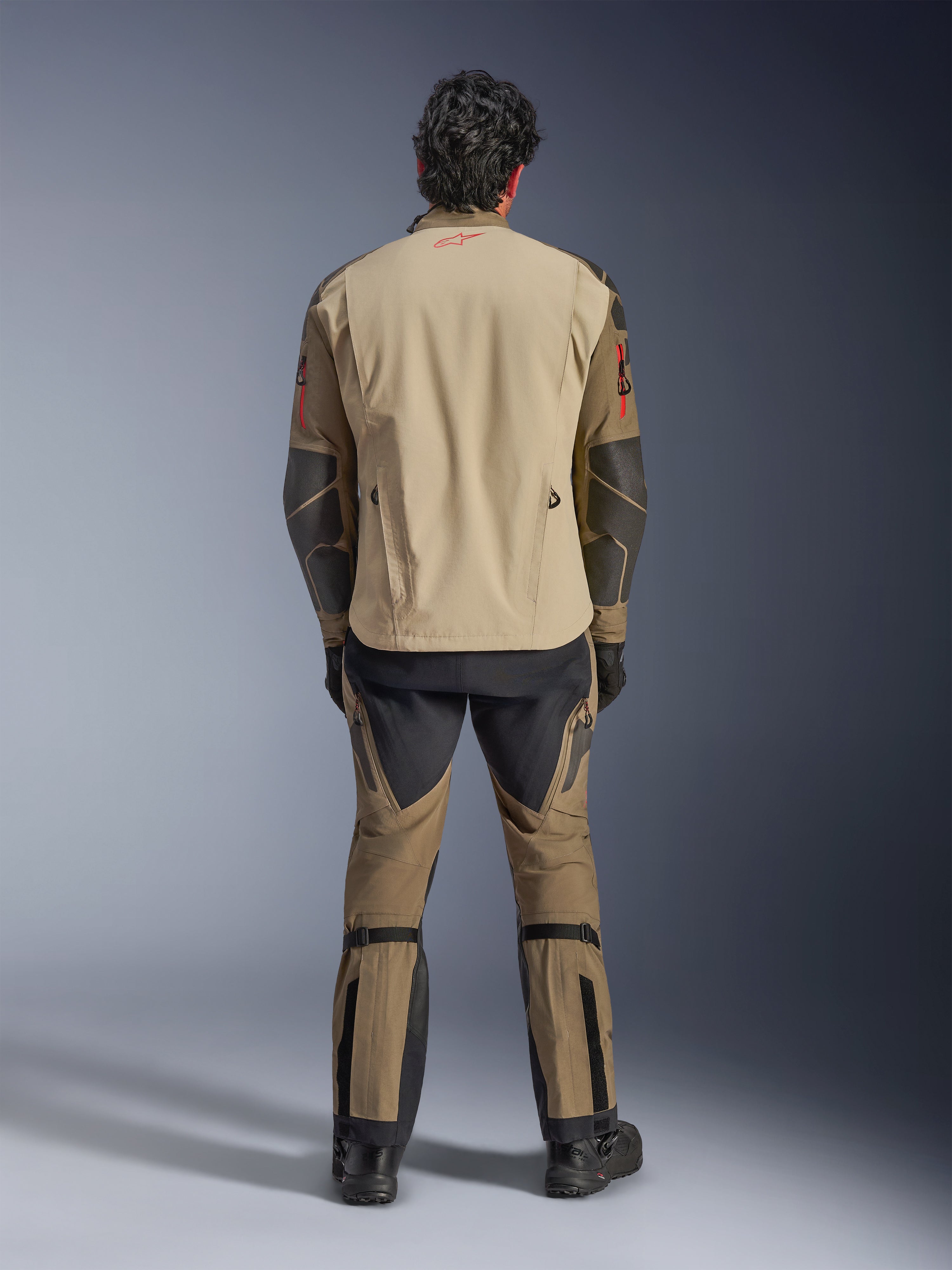 Kurtka Alpinestars Nazca 3L GORE-TEX PRO w kolorze Walnut Rock Brown, widok z tyłu na motocyklistę, widoczne wzmocnienia Superfabric na łokciach i tylne zamki wentylacyjne, noszona z dopasowanymi spodniami adventure, czarnymi butami i rękawicami, wytrzymała techniczna odzież motocyklowa na ekstremalne przygody.