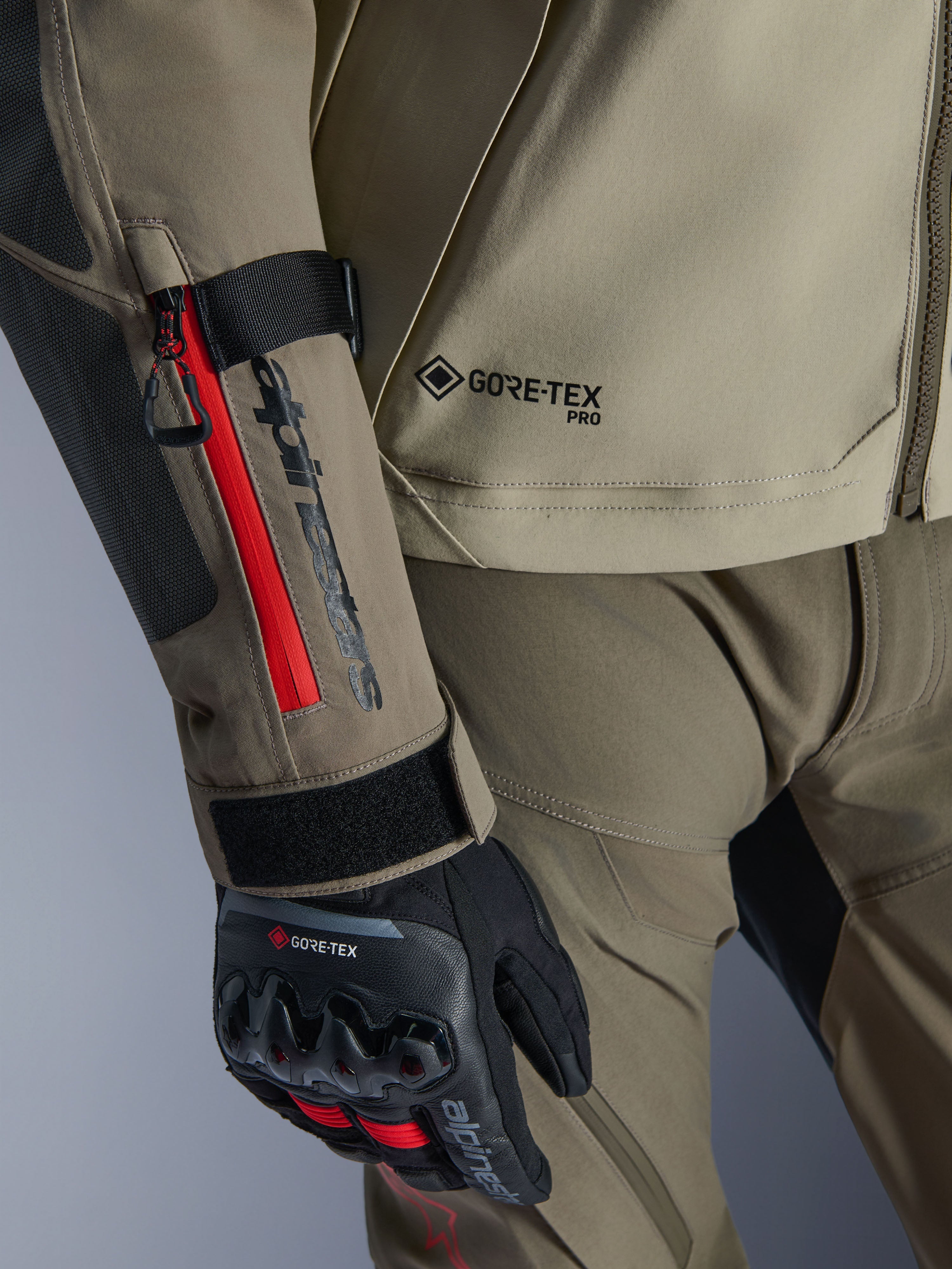 Zbliżenie na kurtkę Alpinestars Nazca 3L GORE-TEX PRO w kolorze Walnut Rock Brown, czerwone akcenty zamków na rękawie, branding GORE-TEX PRO, zestawiona z czarnymi rękawicami motocyklowymi adventure GORE-TEX