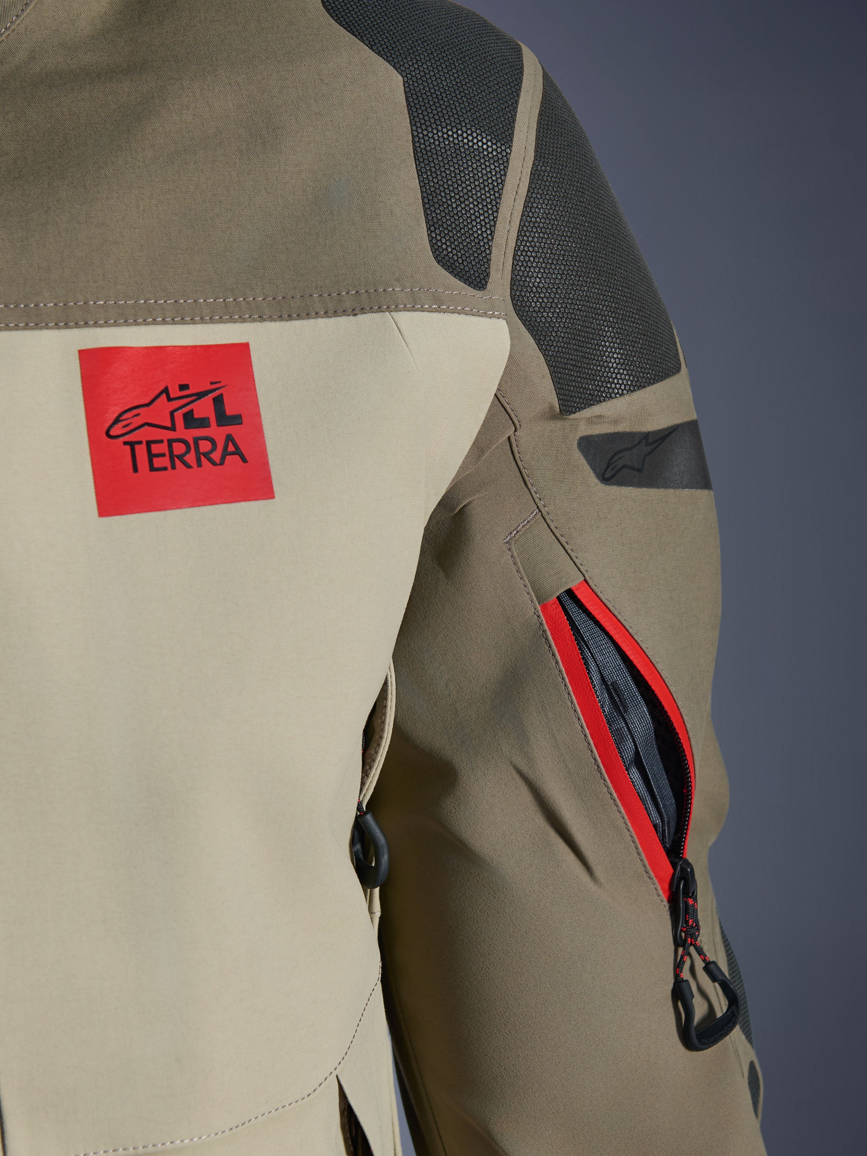 Kurtka Alpinestars Nazca 3L GORE-TEX PRO, Walnut Rock Brown, detal tylnej części ramienia i rękawa, czerwona naszywka z logo „ALL TERRA”, czarne panele Superfabric, zapinany otwór wentylacyjny z czerwonym wykończeniem, odzież motocyklowa adventure