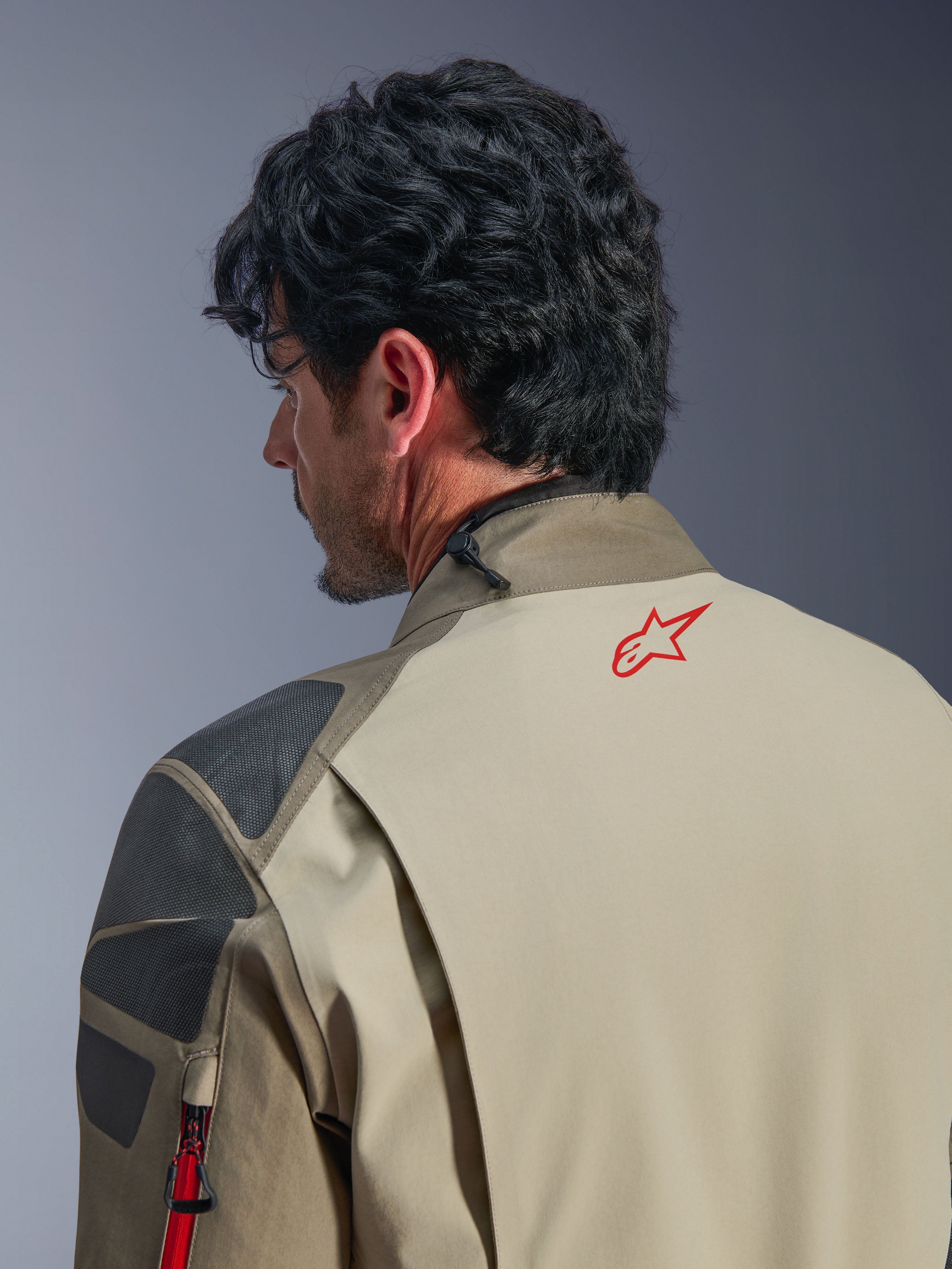 Kurtka Alpinestars Nazca 3L GORE-TEX PRO, kurtka przygodowa w kolorze Walnut Rock Brown, zbliżenie na plecy i ramię, czerwone logo gwiazdy na karku, szare teksturowane panele wzmacniające, wytrzymała konstrukcja tekstylna do jazdy motocyklem