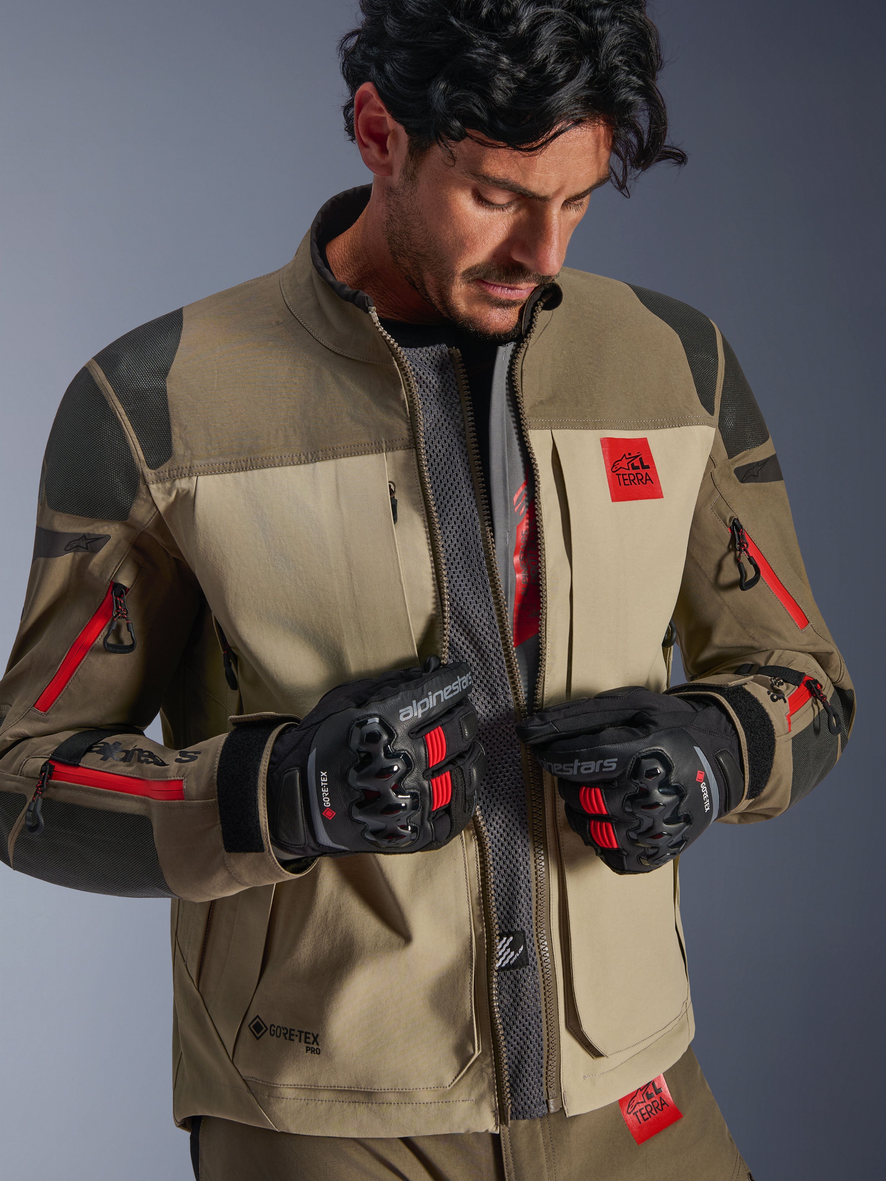 Kurtka Alpinestars Nazca 3L GORE-TEX PRO, Walnut Rock Brown, motocyklowa kurtka adventure z rozpiętym przodem odsłaniającym centralny siatkowy panel wentylacyjny, czerwone logo All Terra, noszona z czarnymi rękawicami motocyklowymi GORE-TEX, odzież techniczna do turystyki off-road