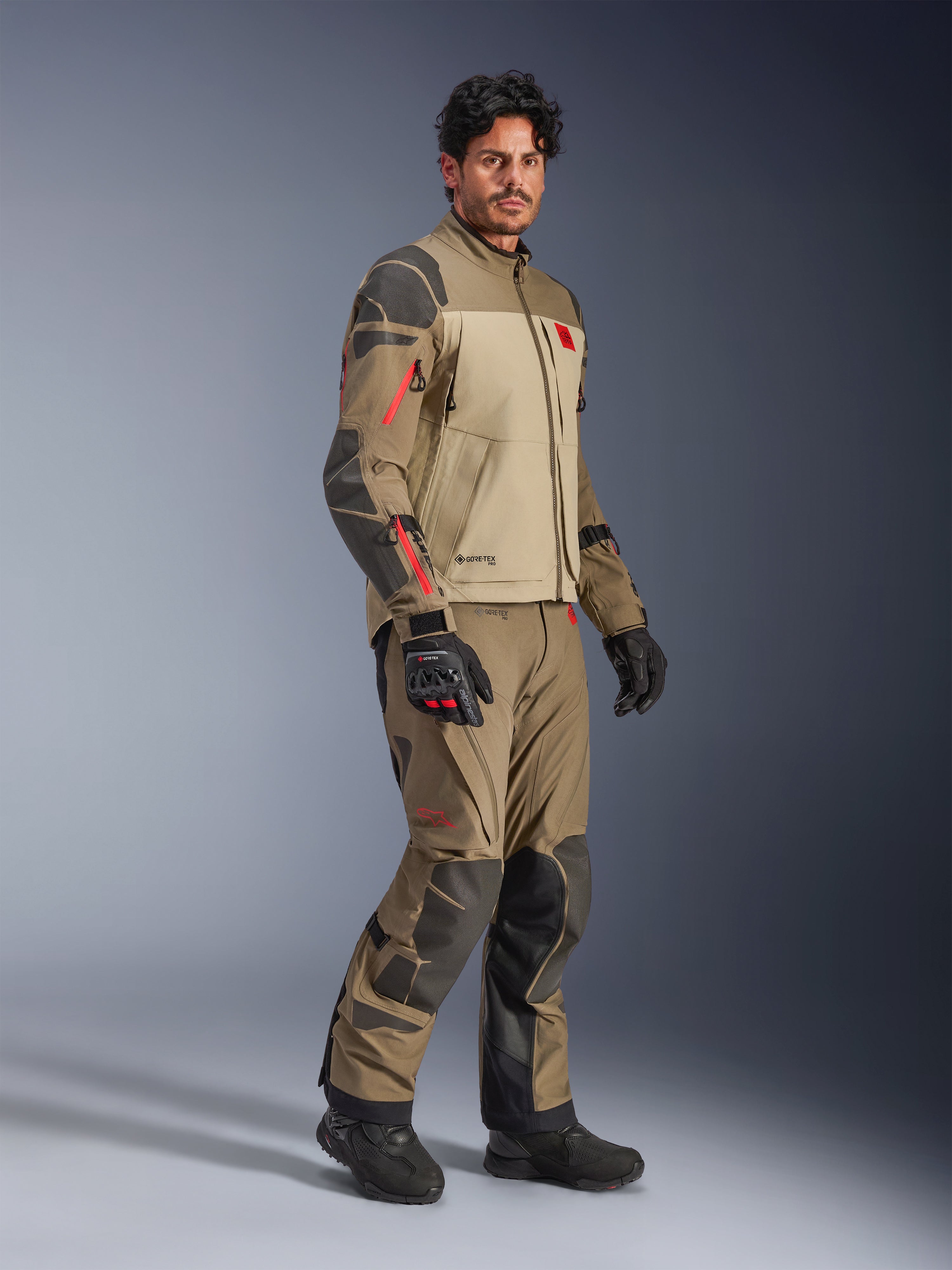 Kurtka Alpinestars Nazca 3L GORE-TEX PRO, Walnut Rock Brown, motocyklowa kurtka przygodowa noszona z pasującymi spodniami i czarnymi rękawicami, z ciemnoszarymi wzmocnieniami, czerwonymi detalami zamków i wysokowydajną laminowaną powłoką.