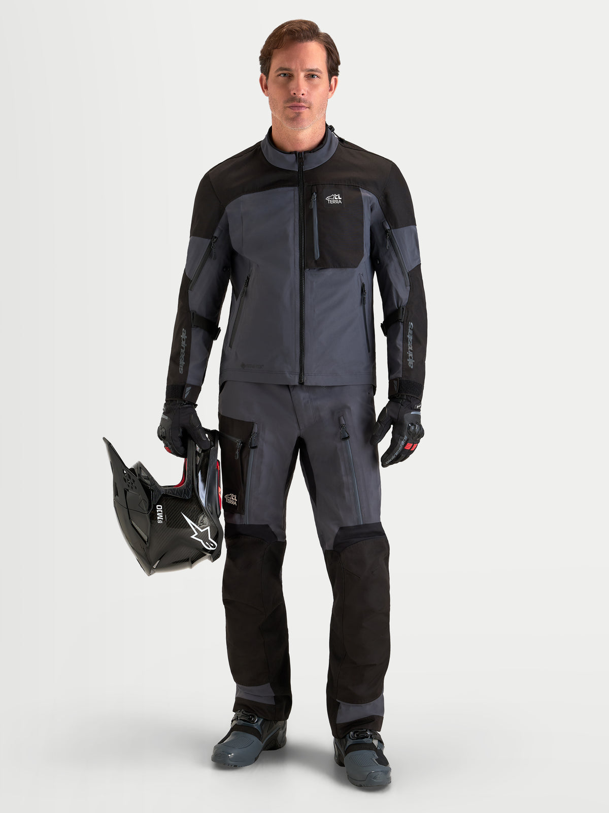 Alpinestars Arenis Gore-Tex Gear Set, męski zestaw motocyklowy adventure kurtka i spodnie w kolorze czarnym i ciemnoszarym, pełny widok z przodu na modela stojącego z kaskiem w ręku w wodoodpornej odzieży tekstylnej