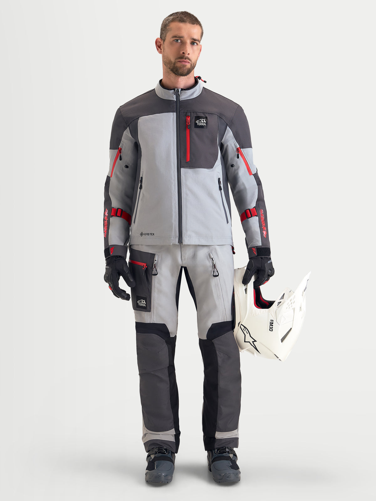 Alpinestars Arenis Gore-Tex Gear Set, męski zestaw odzieży motocyklowej adventure w kolorze szarym i ciemnoszarym z czerwonymi akcentami, widok z przodu na modelu w pasującym stroju, rękawicach i trzymającym biały kask, wodoodporna odzież terenowa