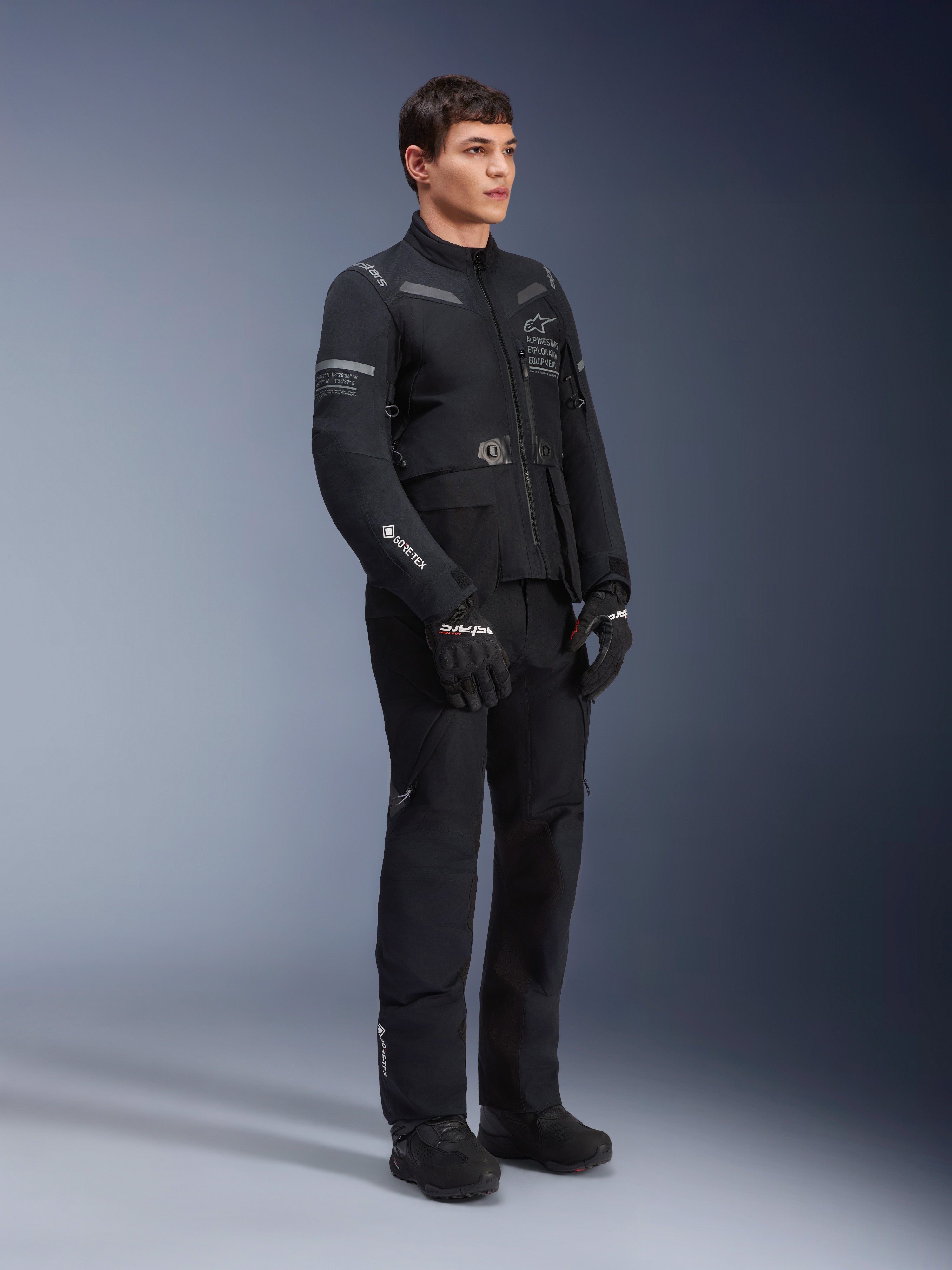 Męski model w kurtce Alpinestars ST-7 2L GORE-TEX w kolorze czarno-ciemnoszarym, motocyklowa kurtka turystyczna adventure, pasujące czarne spodnie i rękawice, ujęcie 3/4 całej sylwetki, widoczne logo GORE-TEX i oznaczenia Alpinestars Exploration Equipment