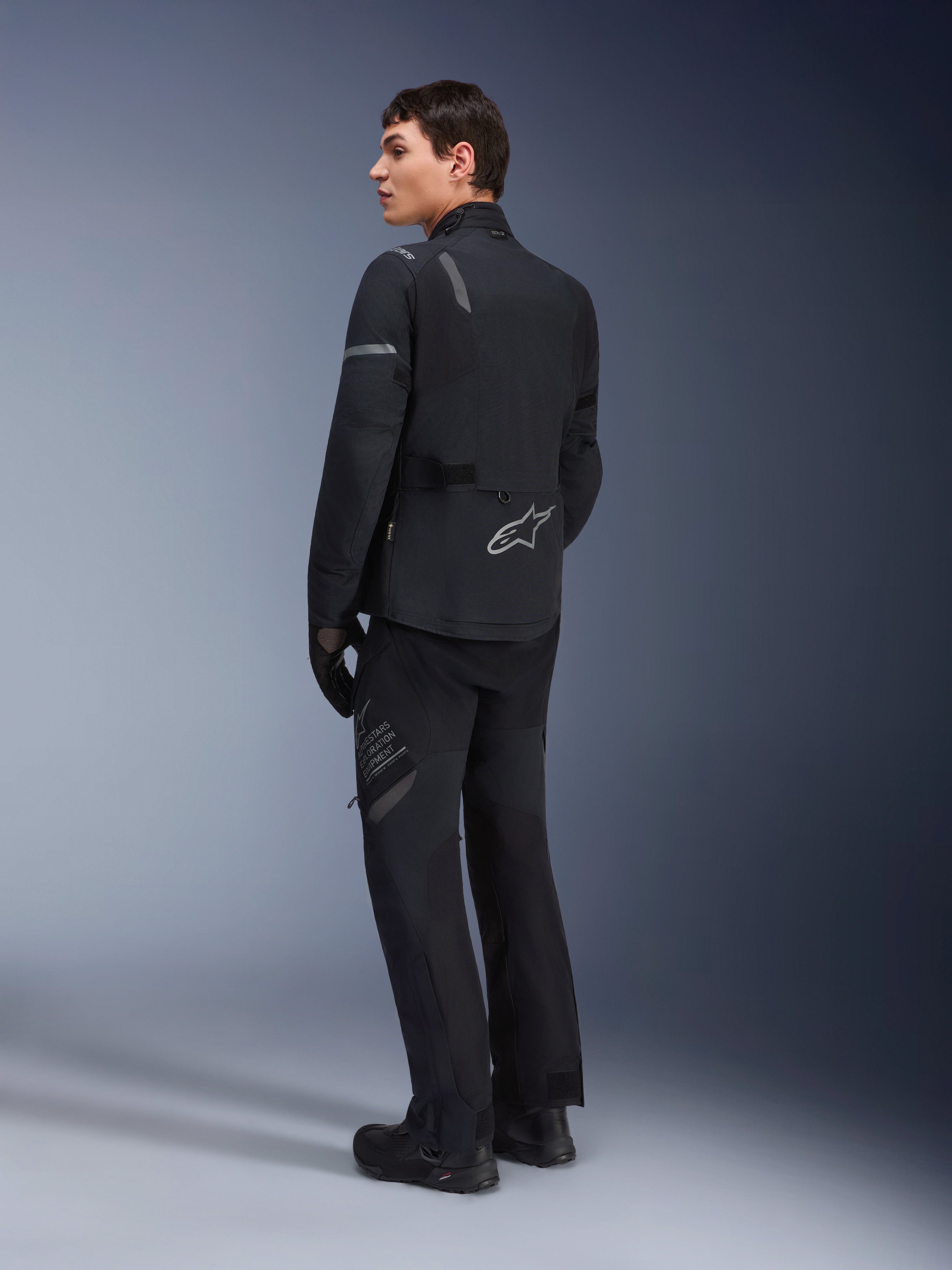 Męski model w kurtce Alpinestars ST-7 2L GORE-TEX w kolorze Black Dark Gray, widok z tyłu pod kątem trzech czwartych ukazujący odblaskowe akcenty i srebrne logo na dole pleców, motocyklowa odzież adventure touring, w zestawie z dopasowanymi czarnymi spodniami i butami