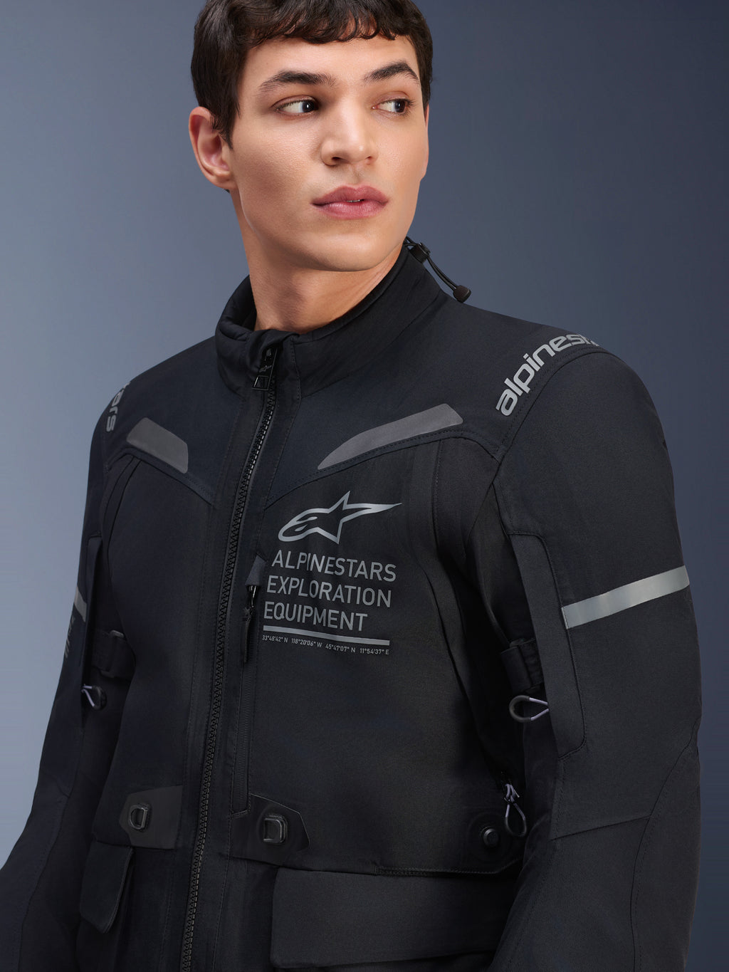 Kurtka Alpinestars ST-7 2L GORE-TEX, kurtka motocyklowa, czarna i ciemnoszara, prezentowana na modelu, z brandingiem Alpinestars Exploration Equipment na klatce piersiowej, odblaskowymi szarymi akcentami i wytrzymałą tekstylną konstrukcją typu adventure