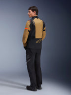 Kurtka Alpinestars ST-7 2L GORE-TEX, motocyklowa kurtka typu adventure, kolor Gobi Brown i czarny, widok z tyłu na modelu ubranym w dopasowane spodnie i buty motocyklowe, z pomarańczowymi akcentami na rękawach, wodoodporną laminowaną powłoką i logo Alpinestars, zaprojektowana do turystyki i jazdy szosowej