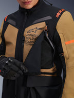 Kurtka Alpinestars ST-7 2L GORE-TEX, motocyklowa kurtka turystyczna adventure, kolor brązowy Gobi Brown i czarny, zbliżenie na rozpięty panel wentylacyjny na klatce piersiowej odsłaniający oddychającą siateczkę, z brandingiem Alpinestars Exploration Equipment i pomarańczowymi akcentami.