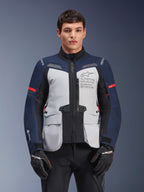 Kurtka Alpinestars ST-7 2L GORE-TEX, motocyklowa kurtka turystyczna adventure, lodowoszara i ciemnoniebieska z czerwonymi akcentami, wodoodporna i oddychająca, prezentowana na modelu w czarnych rękawicach, z logo Alpinestars Exploration Equipment na klatce piersiowej