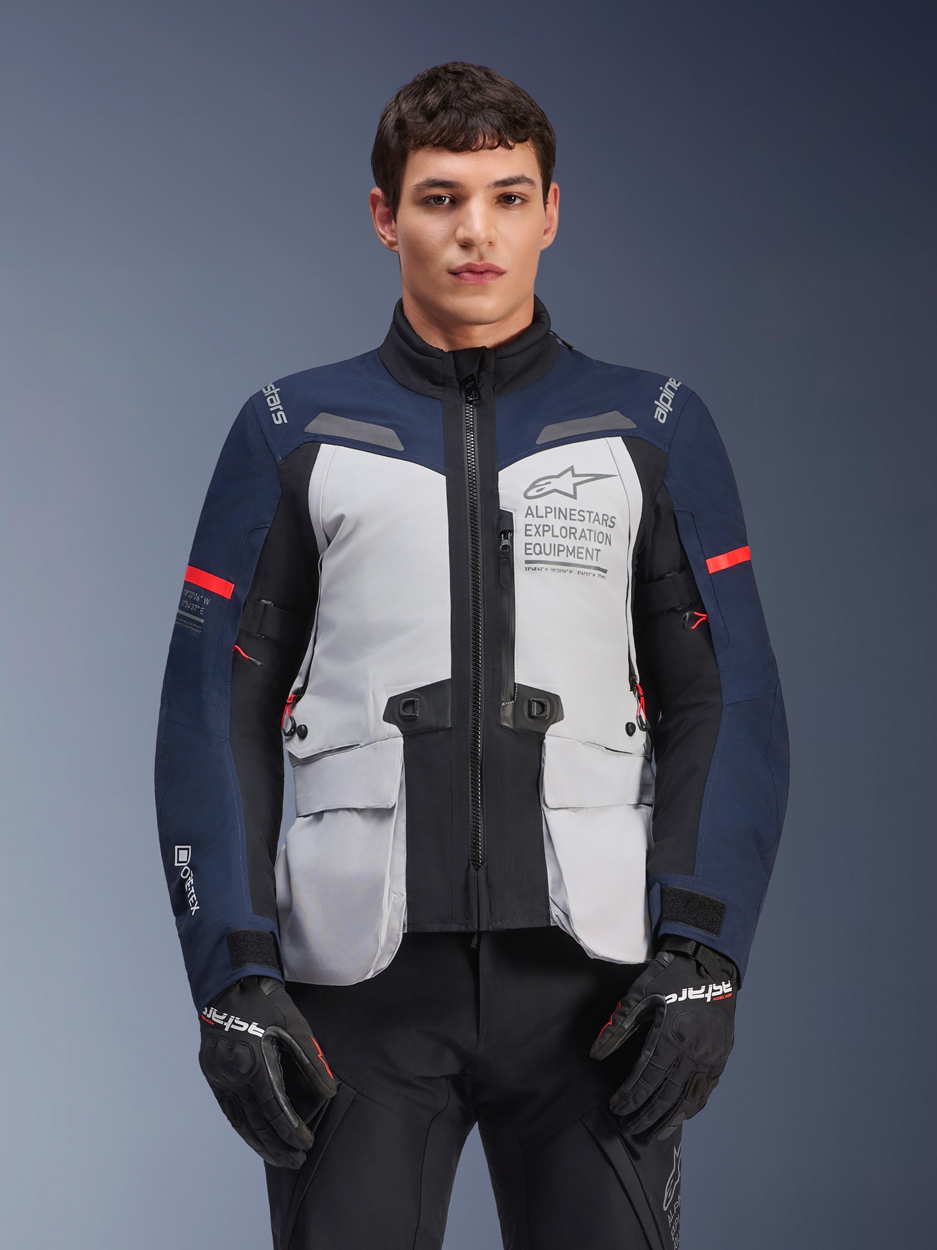 Kurtka Alpinestars ST-7 2L GORE-TEX, motocyklowa kurtka turystyczna adventure, lodowoszara i ciemnoniebieska z czerwonymi akcentami, wodoodporna i oddychająca, prezentowana na modelu w czarnych rękawicach, z logo Alpinestars Exploration Equipment na klatce piersiowej